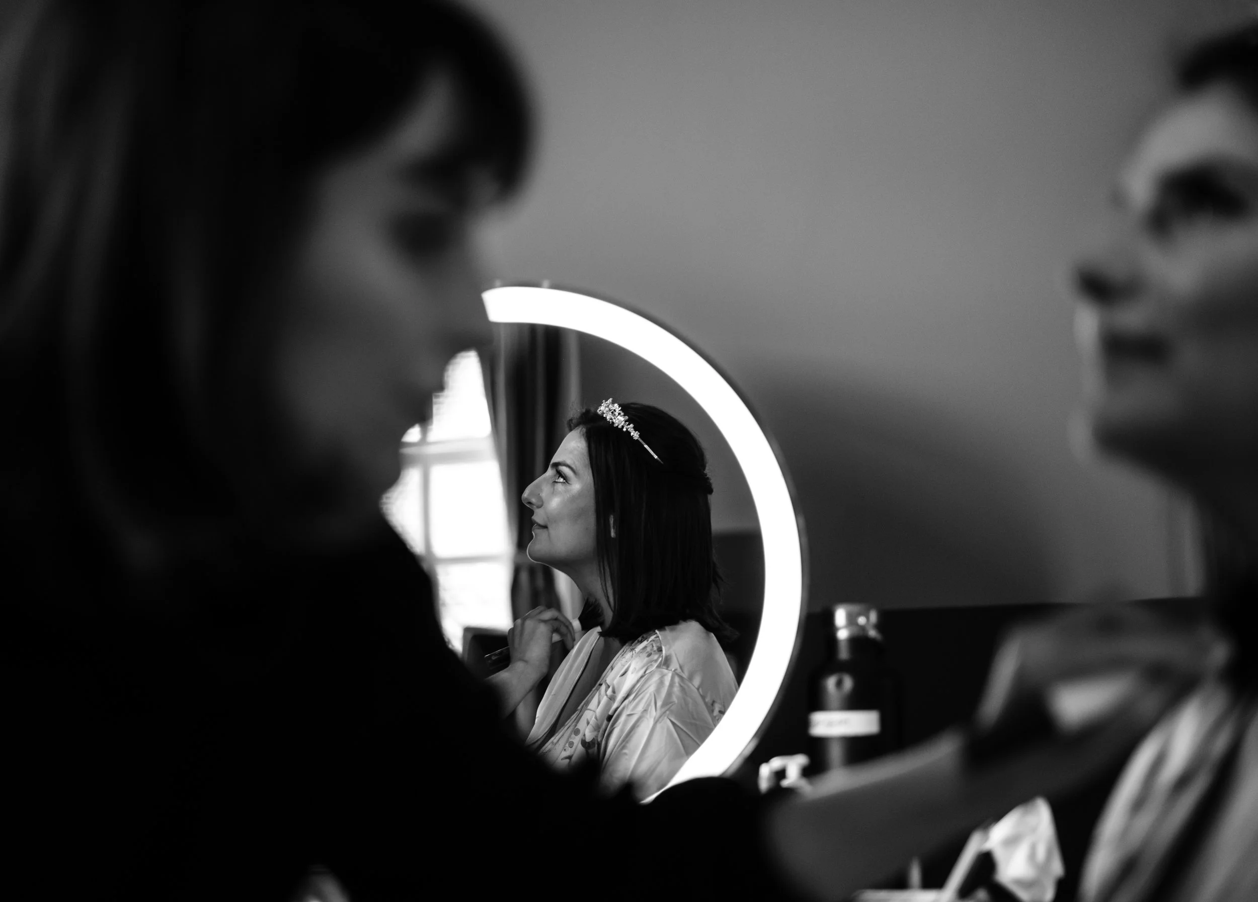 A woman looking into a mirror getting ready, with a stylist adjusting her hair, in a room with a circular lighted mirror.