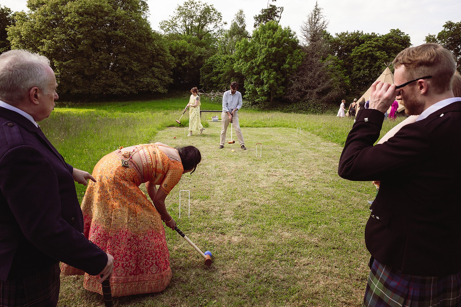 People playing lawn games outdoors, some in traditional clothing, with trees and a tent in the background.