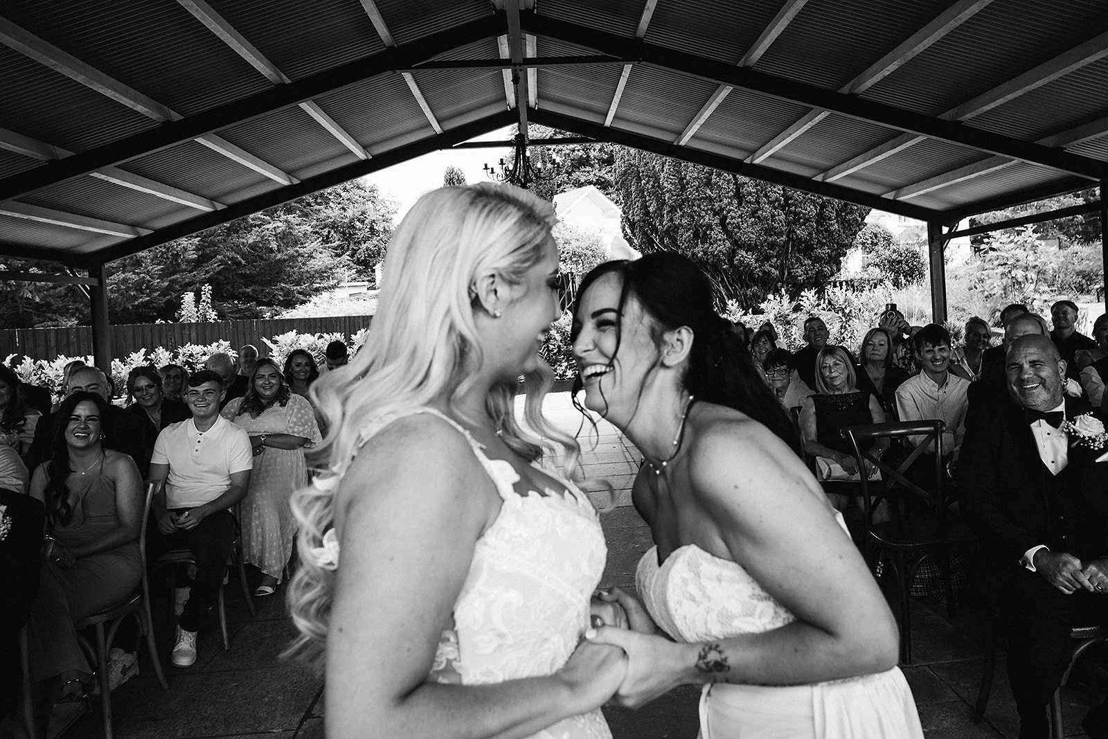 Black and white photo of two women in wedding dresses smiling and holding hands at a wedding reception, surrounded by seated guests under a covered outdoor area.