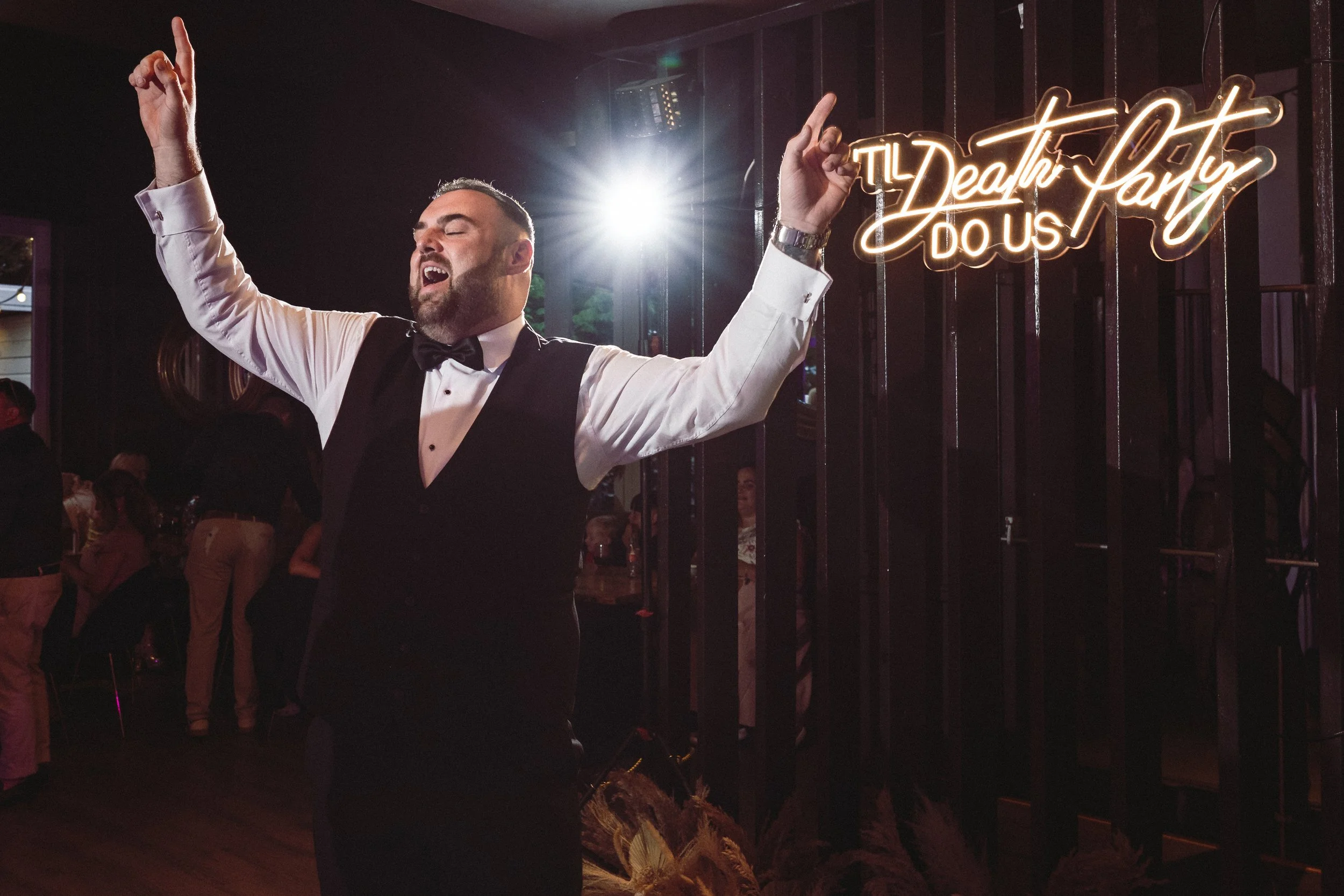 A man in a black tuxedo and bow tie singing passionately with arms raised in a party setting, with a neon sign reading 'The Death Party Do Us' in the background.