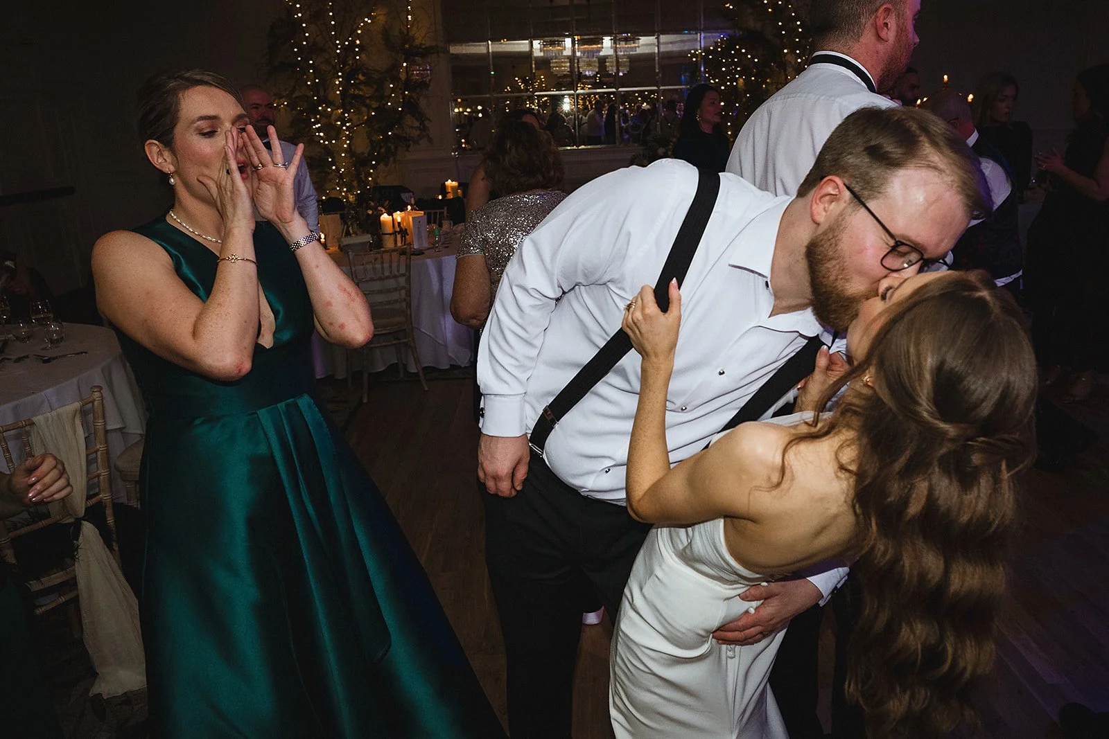 A wedding reception scene where a man and woman are sharing a kiss, a woman in an emerald green dress is reacting with joy, covering her face with her hands.