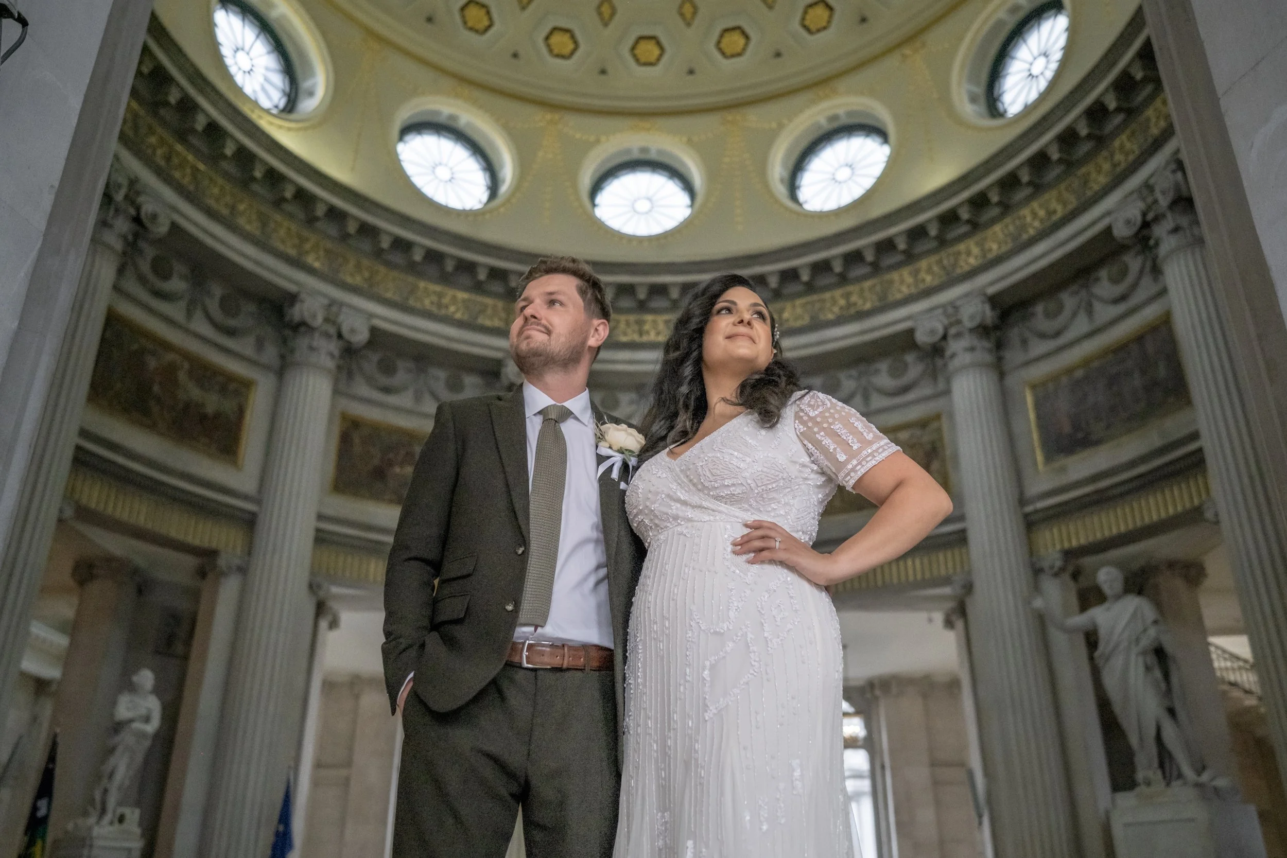 A couple in wedding attire standing inside a grand, domed building with ornate architecture, tall columns, and stained glass windows.