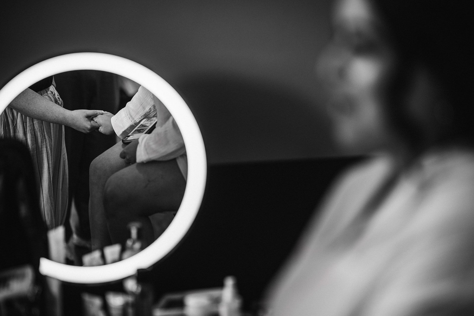 A person sitting in front of a mirror, holding a phone, while another person stands and extends their hand. The mirror reflects the scene, focusing on two people holding hands.