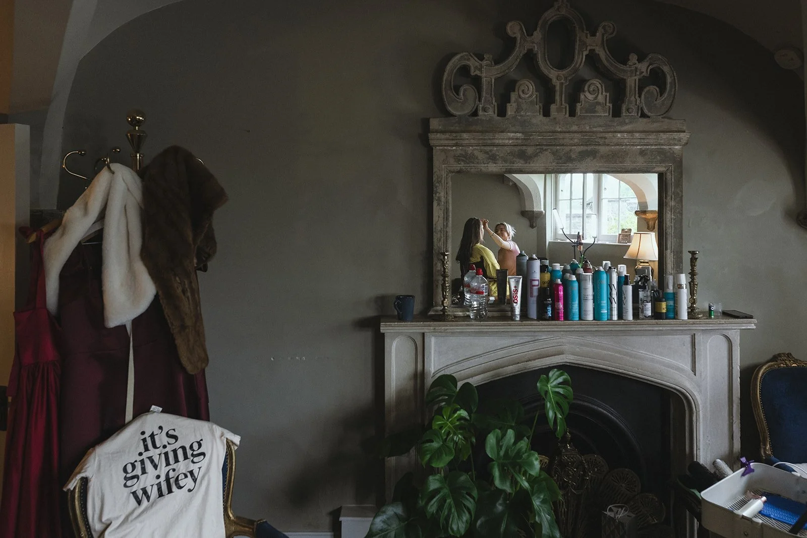 A dark living room with a large mirror above a fireplace, cluttered with hair and makeup products. To the left, a coat rack with a fur-lined coat, and a chair with a cloth that reads 'it's giving wifey.' A woman is getting ready in the mirror, receiv