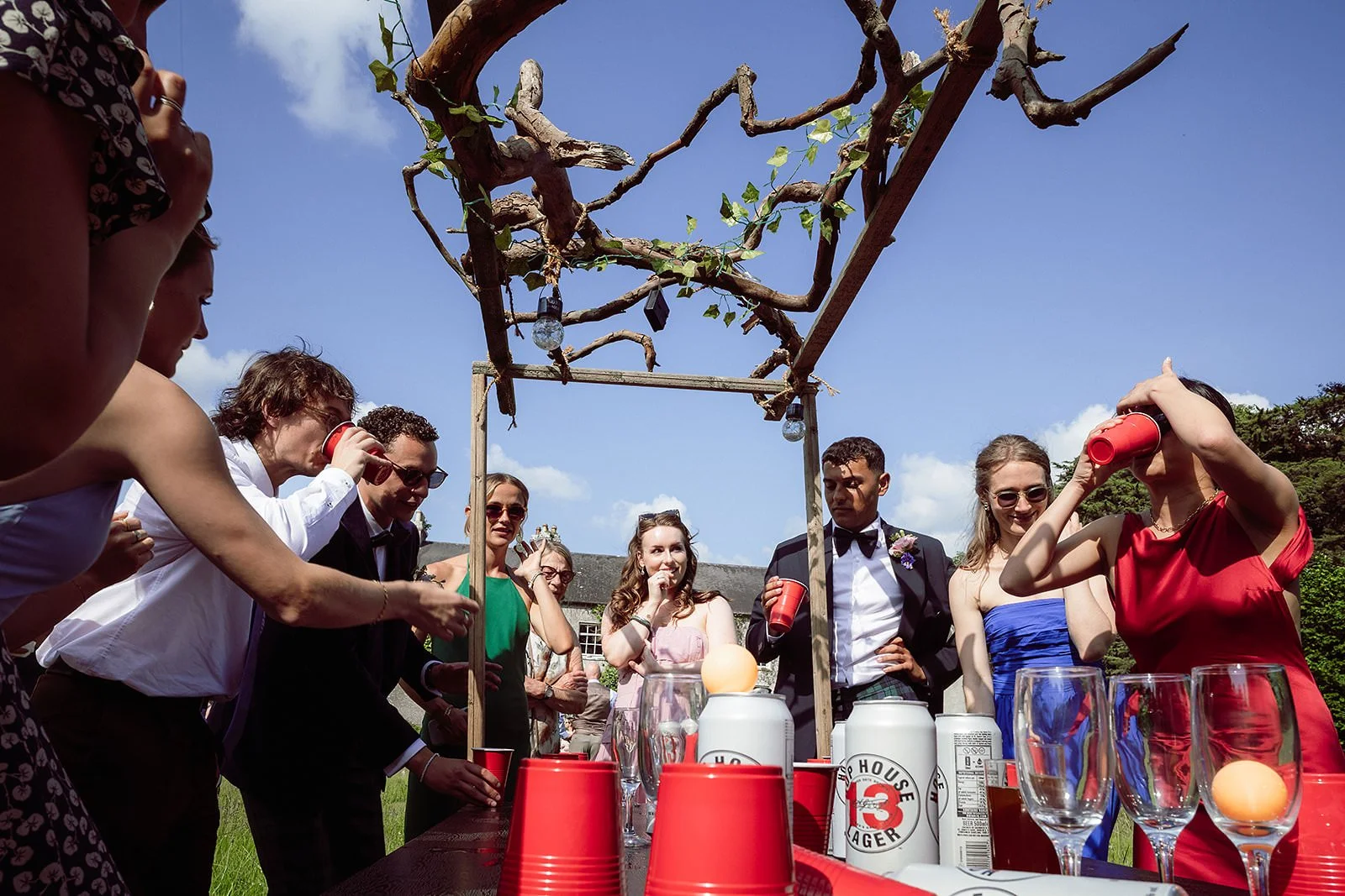 A group of people dressed in formal attire around a table outdoors with a DIY wooden structure and tree branches overhead, preparing for a drink activity during a celebration or party.