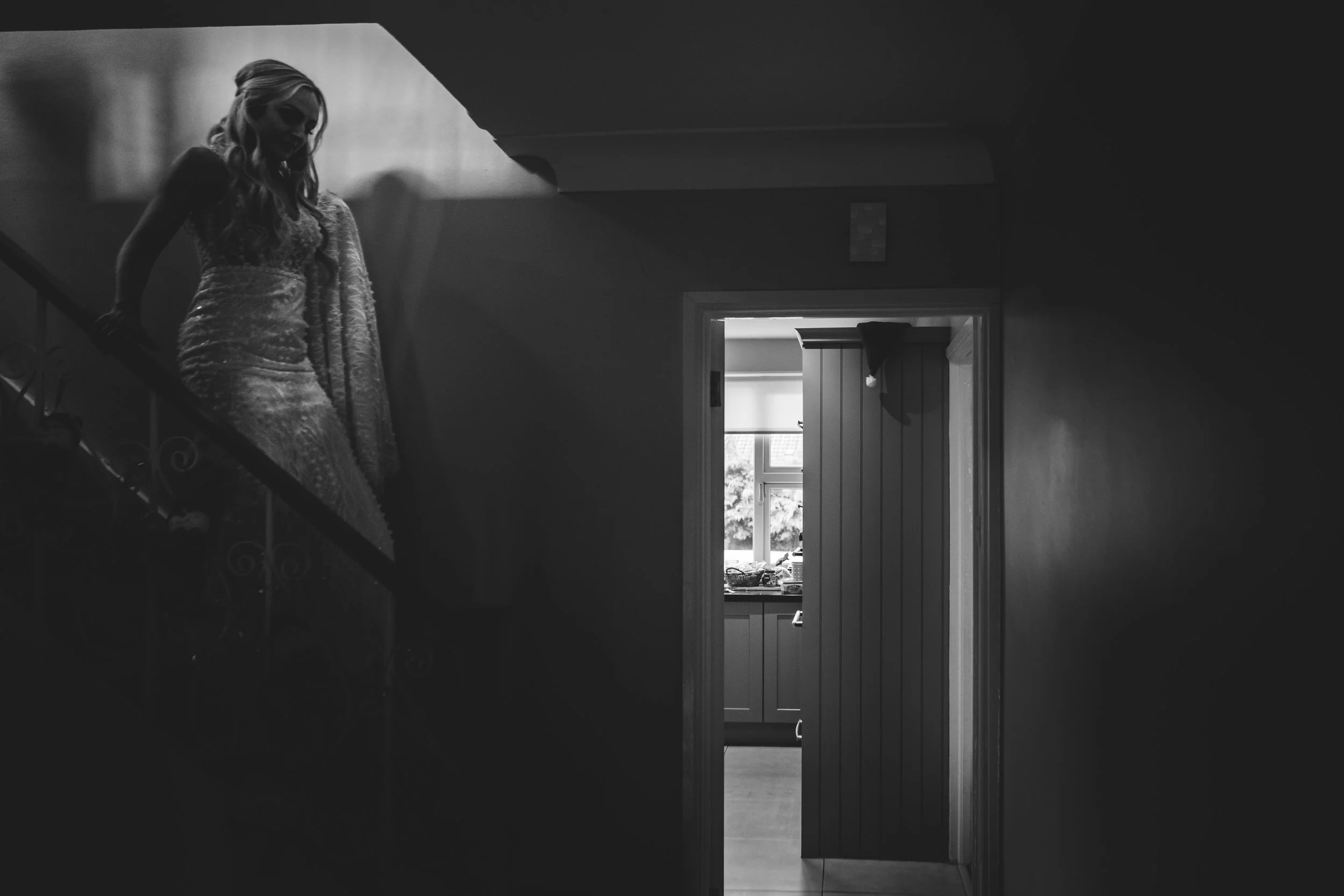 A woman in a patterned dress standing on a staircase, with a domestic scene visible through a doorway in a black and white photo.