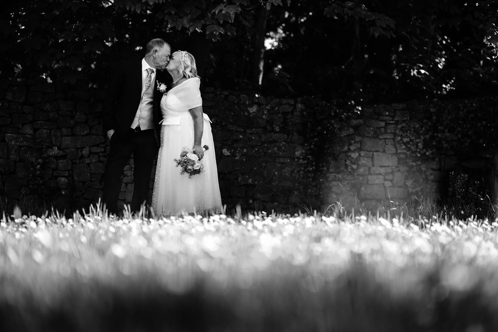 Black and white photo of an elderly man and woman sharing a kiss, with the woman holding a bouquet of roses, standing outdoors in front of a stone wall and surrounded by trees.