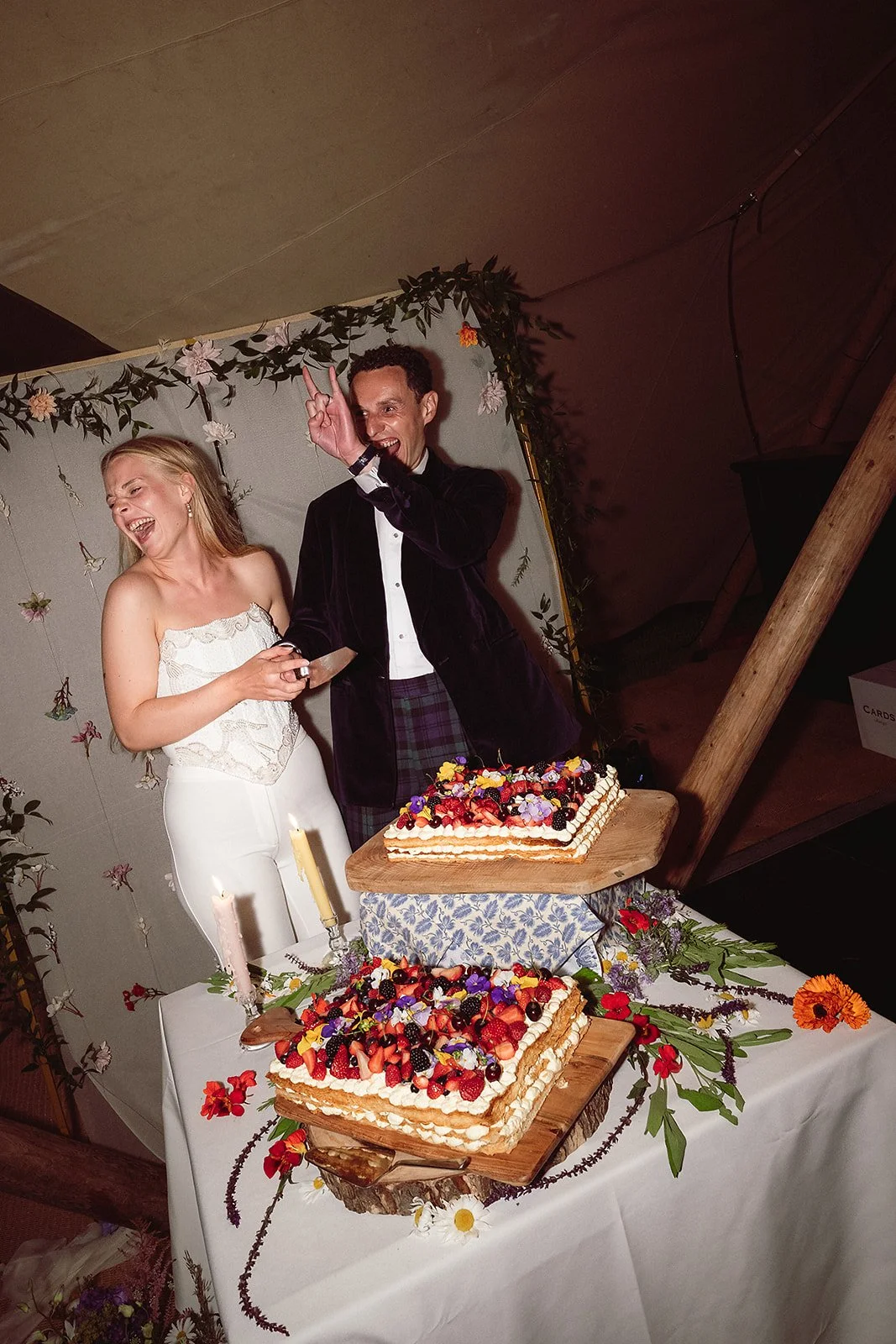A couple celebrating with two large decorated cakes on a table, surrounded by candles and flowers, at an indoor event.