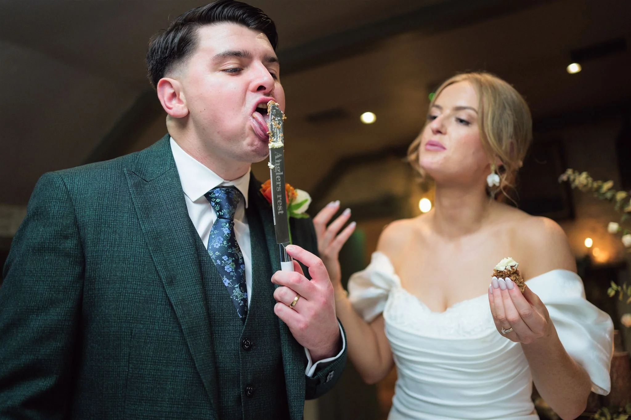 A man in a suit with a blue floral tie is feeding himself wedding cake with a utensil, while a woman in a white off-shoulder wedding dress looks on, holding a piece of cake. The background is warmly lit, indicating they are at a wedding reception.