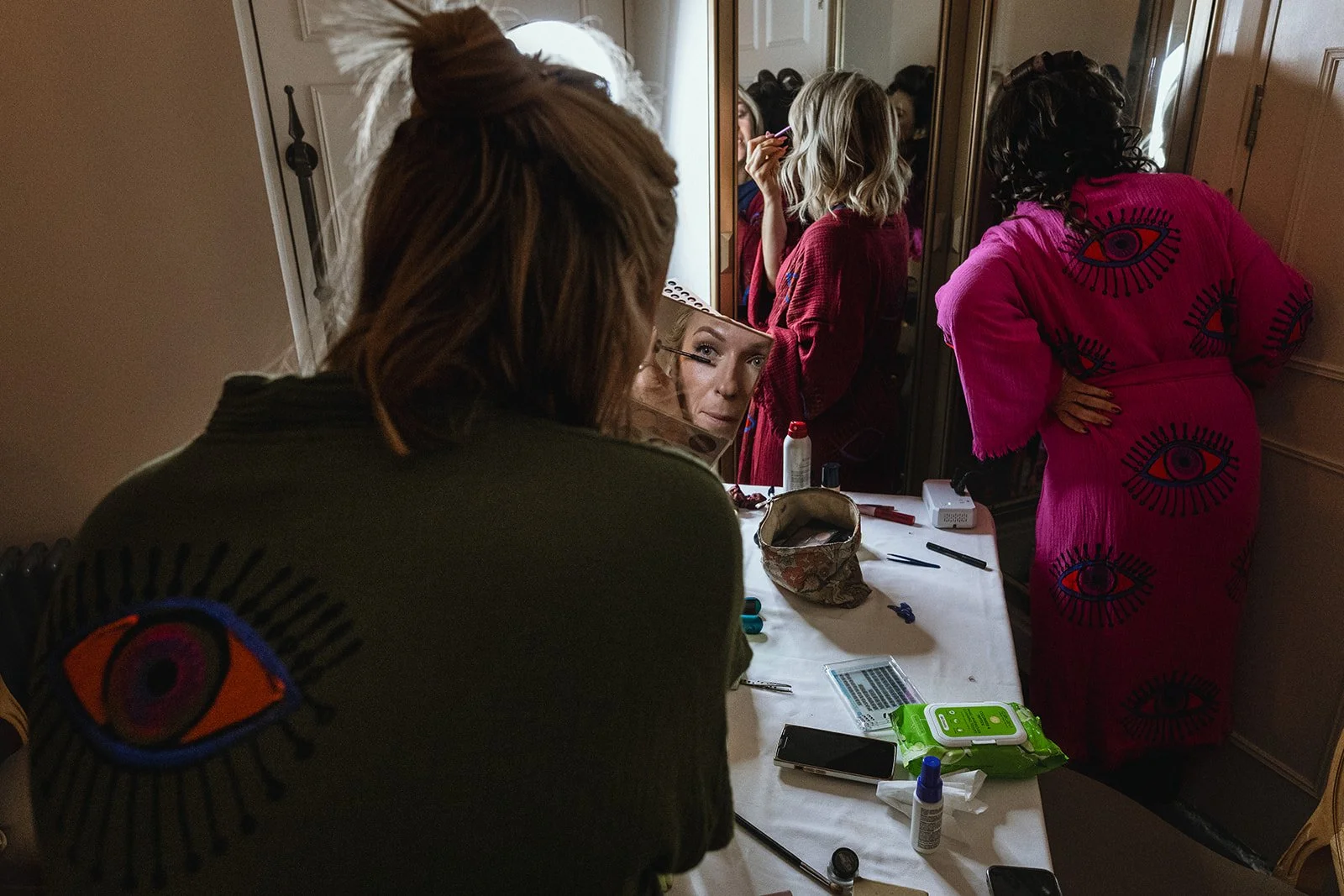 A woman with shoulder-length blonde hair is looking in a mirror while applying makeup. She is wearing a red robe. The mirror reflects her face and part of a woman with curly hair in a pink dress with eye and eyelash patterns, who is standing with her