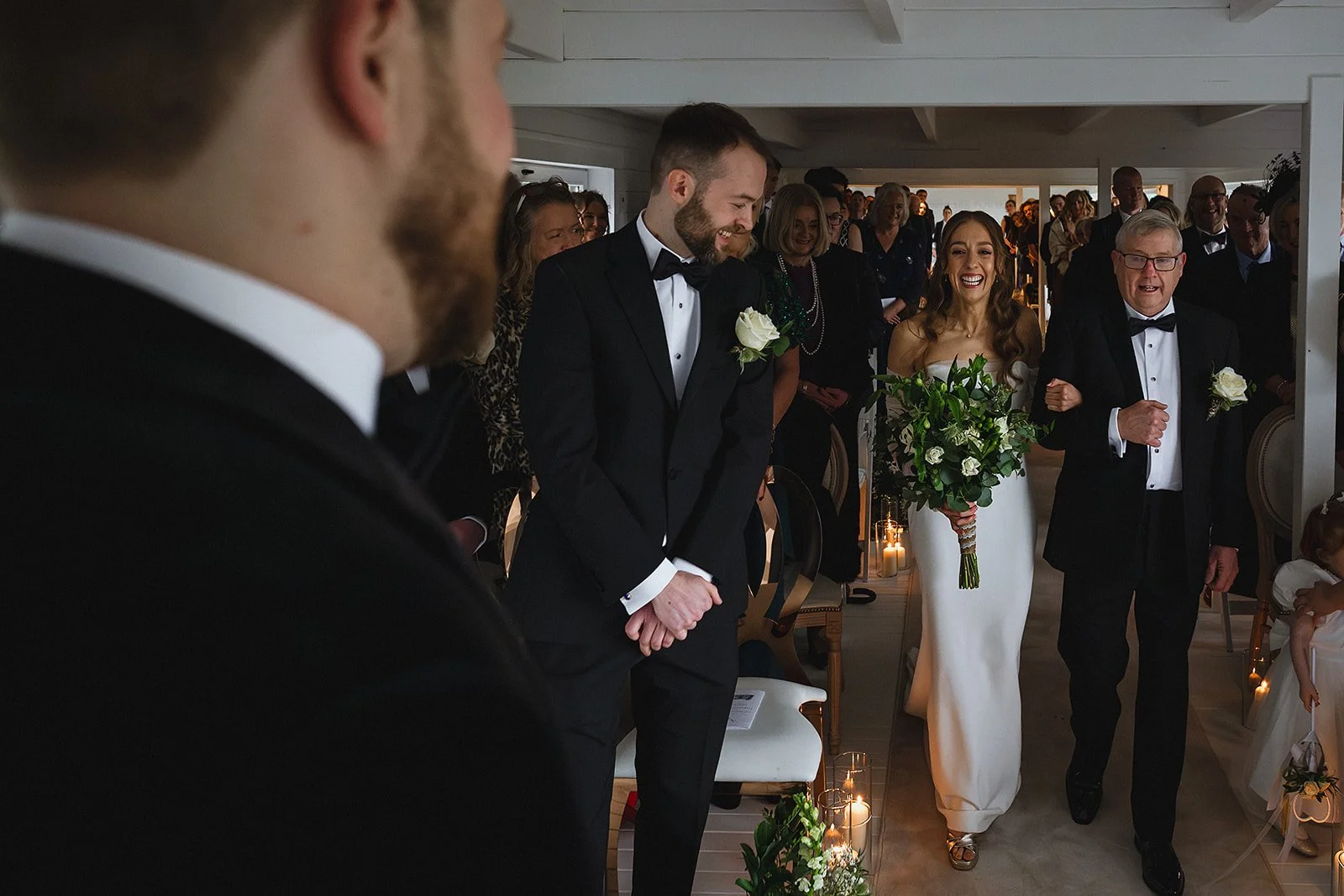 A bride in a white wedding dress walking down an aisle, holding a bouquet, accompanied by an older man in a tuxedo, with guests standing and smiling in the background.