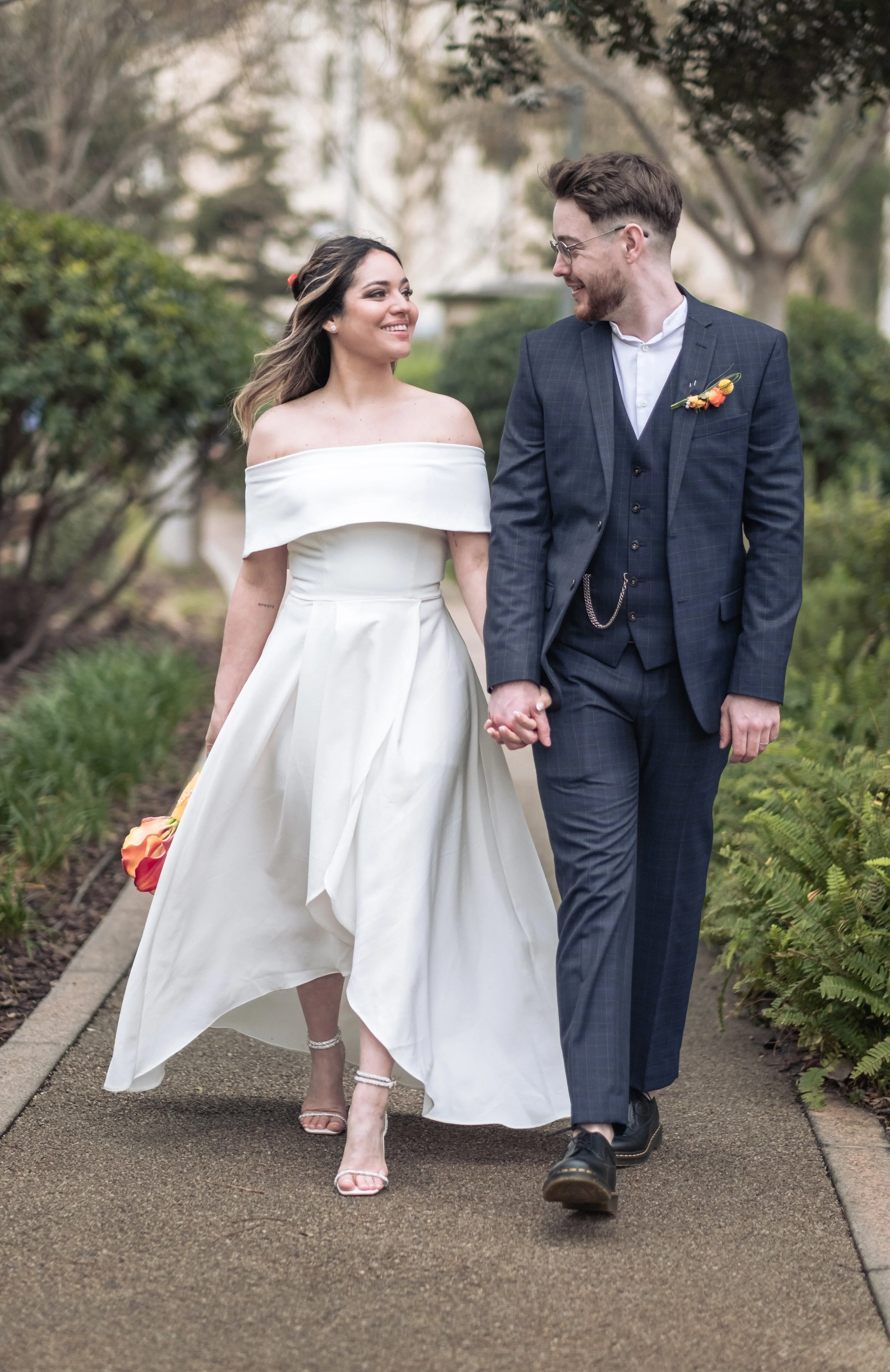 A smiling couple, a bride in a white off-the-shoulder dress and a groom in a dark suit, walk hand-in-hand outdoors on a pathway surrounded by greenery, holding hands and looking at each other.