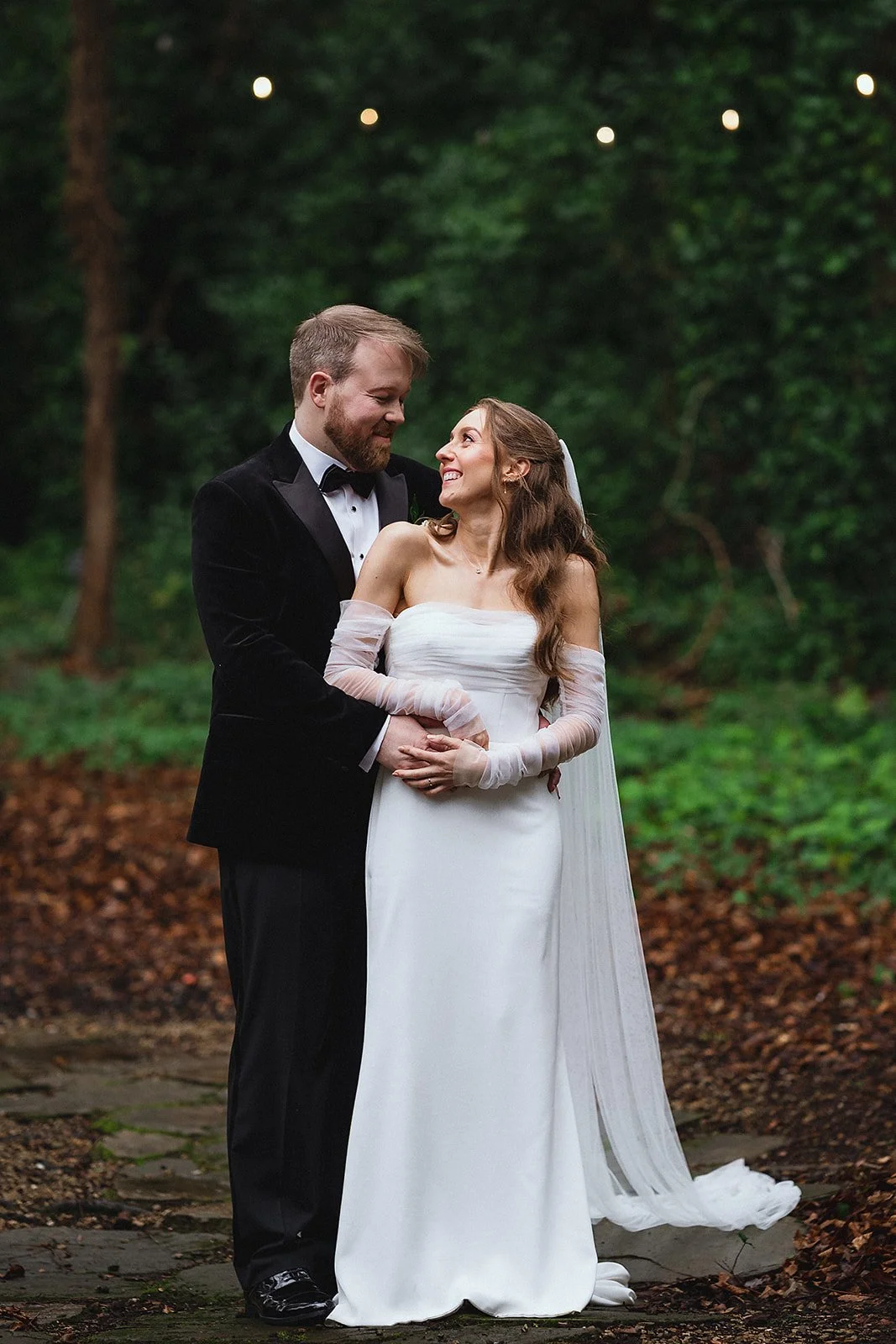 A bride and groom stand together outdoors in a wooded area, smiling and looking at each other. The bride is in a white strapless wedding gown with long sheer gloves, and the groom is in a black tuxedo with a bow tie.