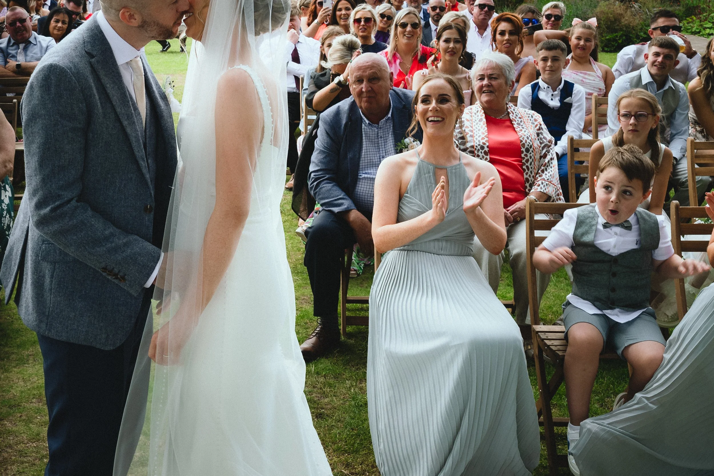 A wedding ceremony outdoors with guests seated on wooden chairs; the bride and groom are standing before the officiant, and guests are smiling and clapping.