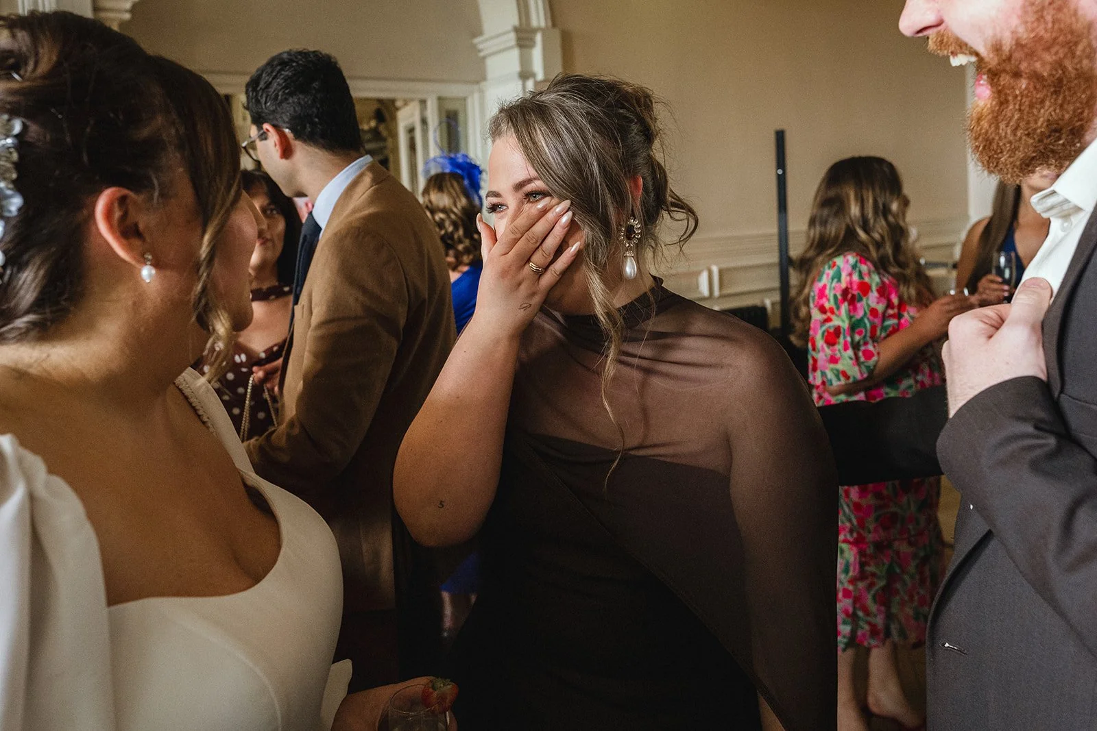A woman in a black dress covering her mouth and smiling at a social event, surrounded by other people in formal attire.