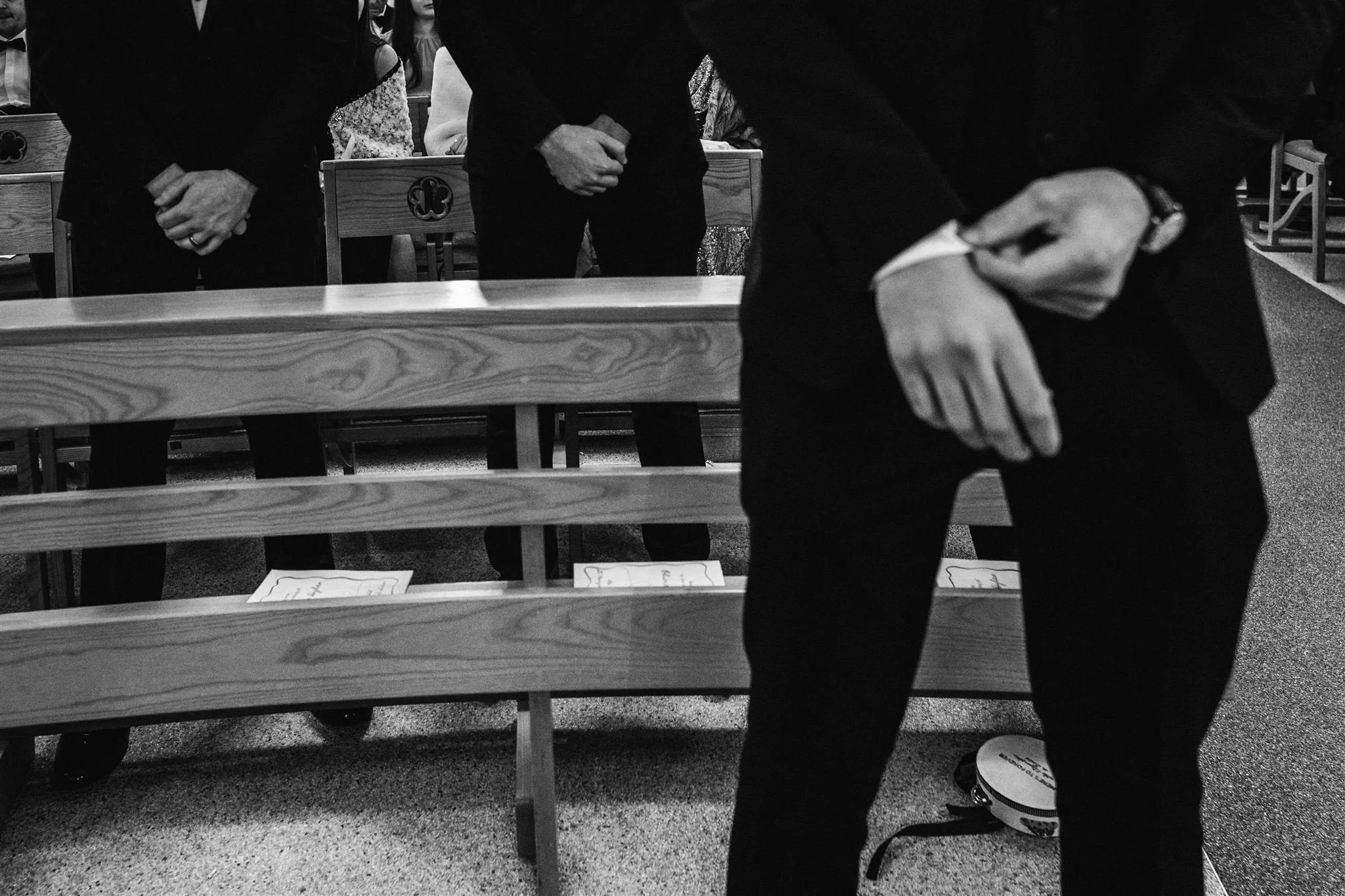 People kneeling and bowing their heads with hands clasped in prayer inside a church, with wooden pews and sheets of paper placed on the pews.