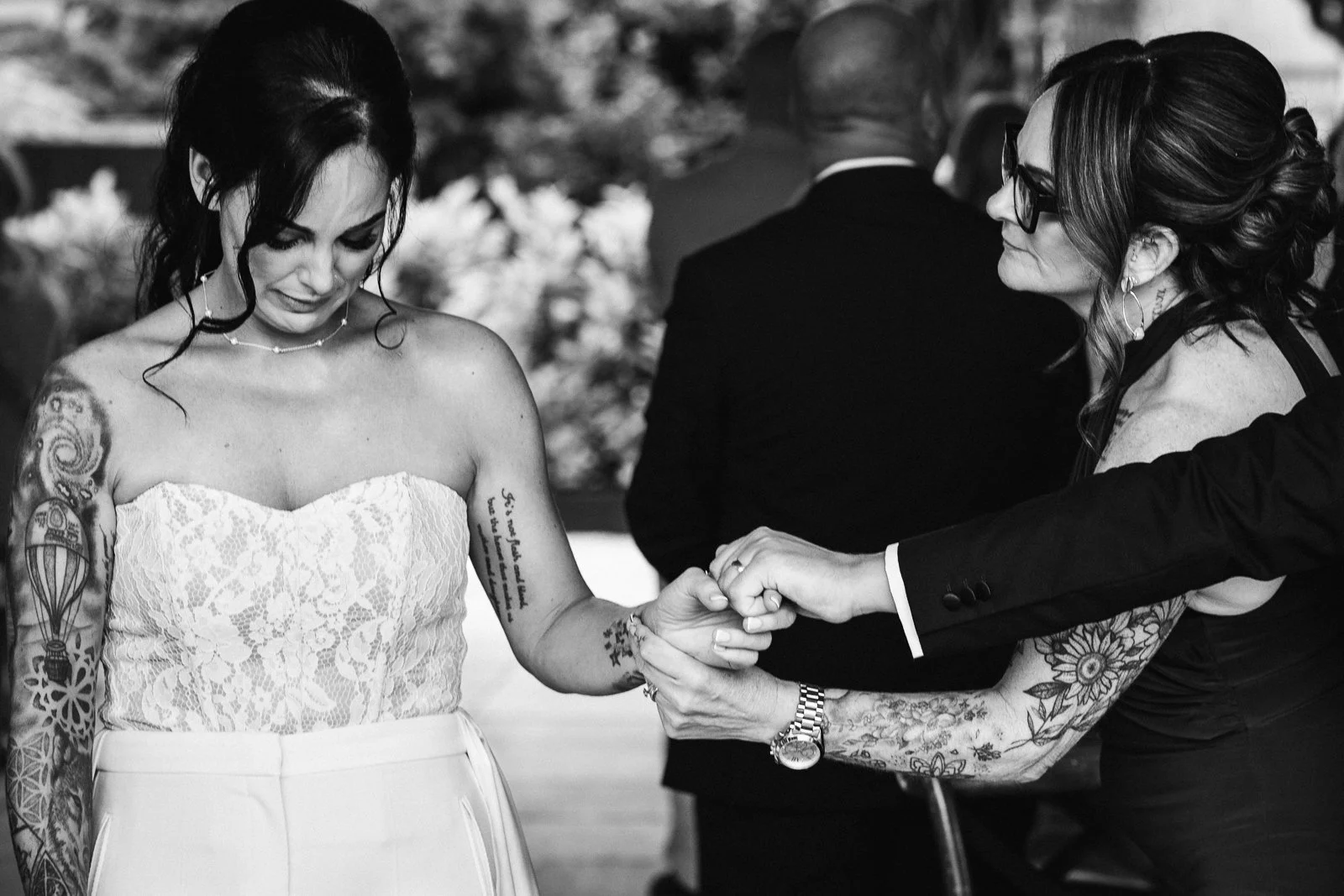 A bride with tattoos on her arms and shoulders, wearing a strapless lace dress, holds hands with another woman who is tattooed and wearing glasses, during a wedding ceremony.