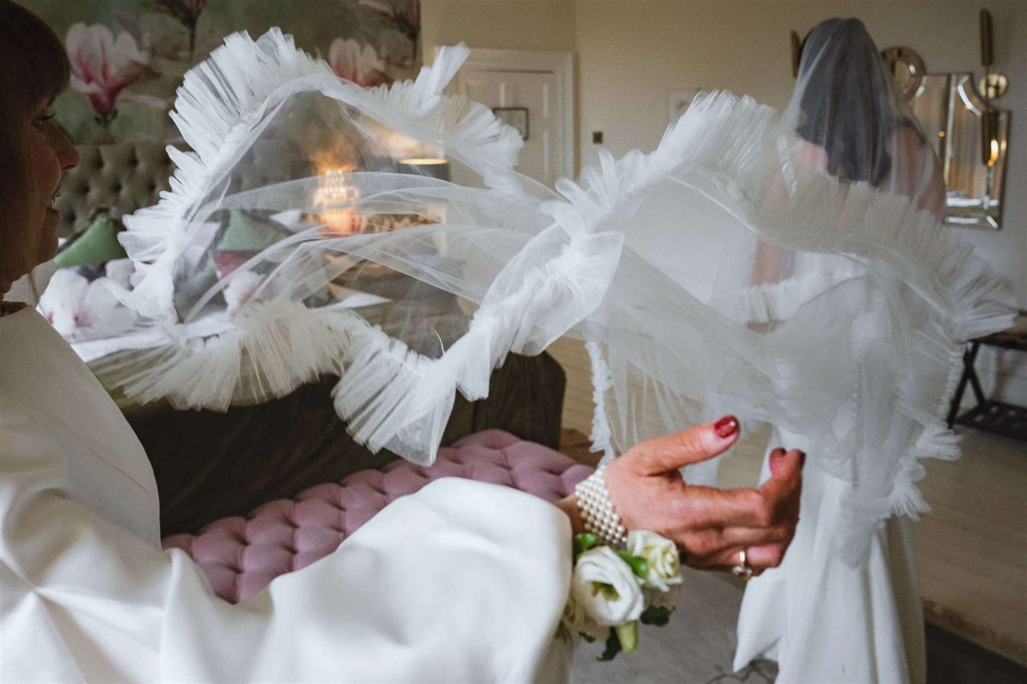 A woman holding a white fishnet wedding veil with ruffled edges, dressed in a white outfit, with a woman in the background and floral decor in a softly lit room.