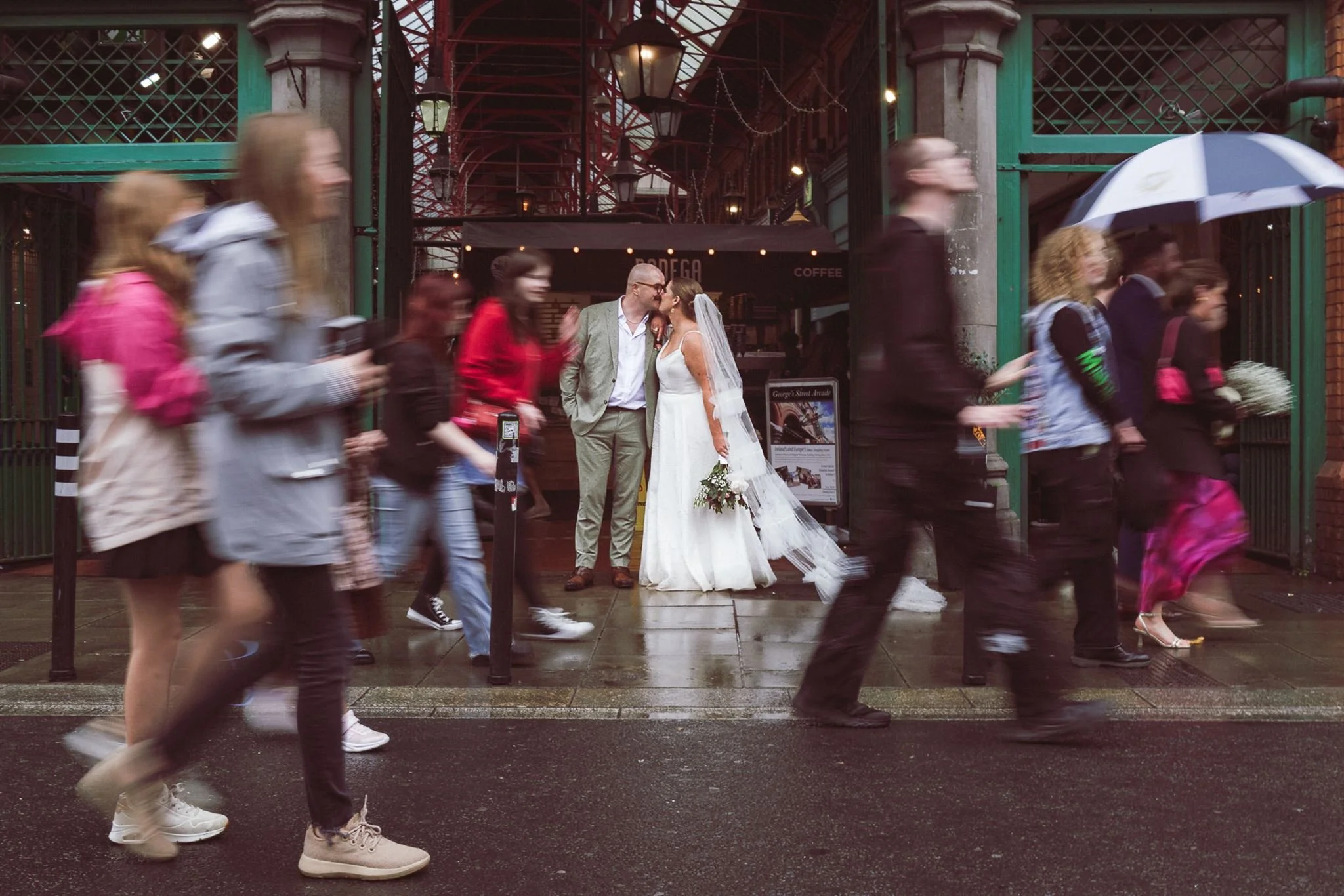 A bride and groom sharing a kiss outside, surrounded by people walking with umbrellas on a rainy day.