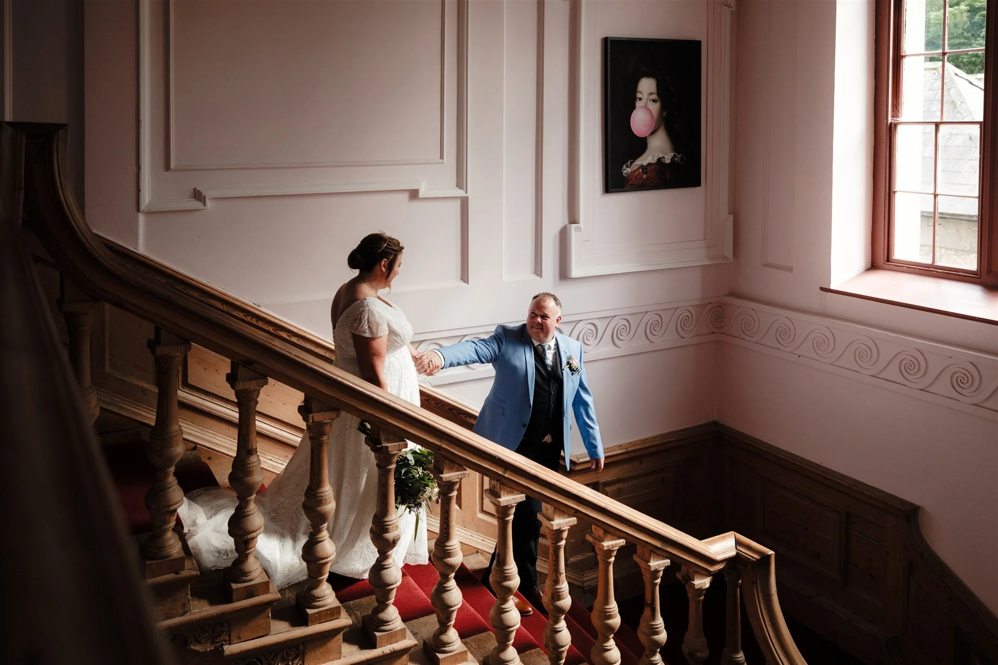 A bride and groom are standing on a staircase inside a building, holding hands and smiling at each other. The bride is wearing a white wedding dress and the groom is in a blue suit. There is a large portrait of a woman with a pink bubblegum bubble on