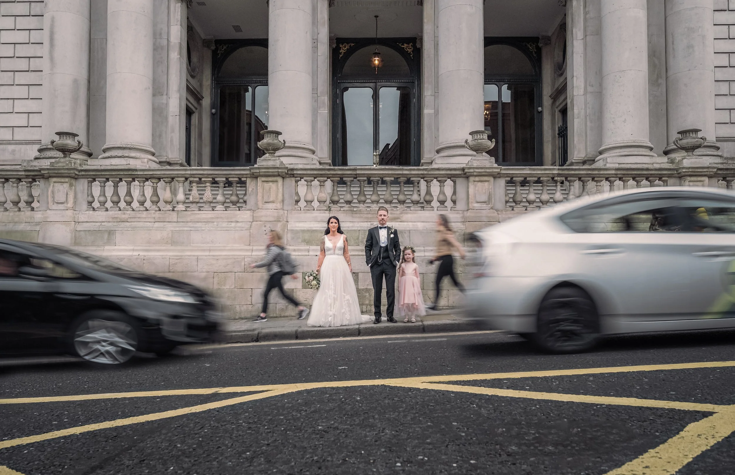 A bride in a white wedding gown, a groom in a dark suit, and a young girl in a light pink dress standing on a city sidewalk in front of a large stone building with columns. Two cars are driving past in the foreground.