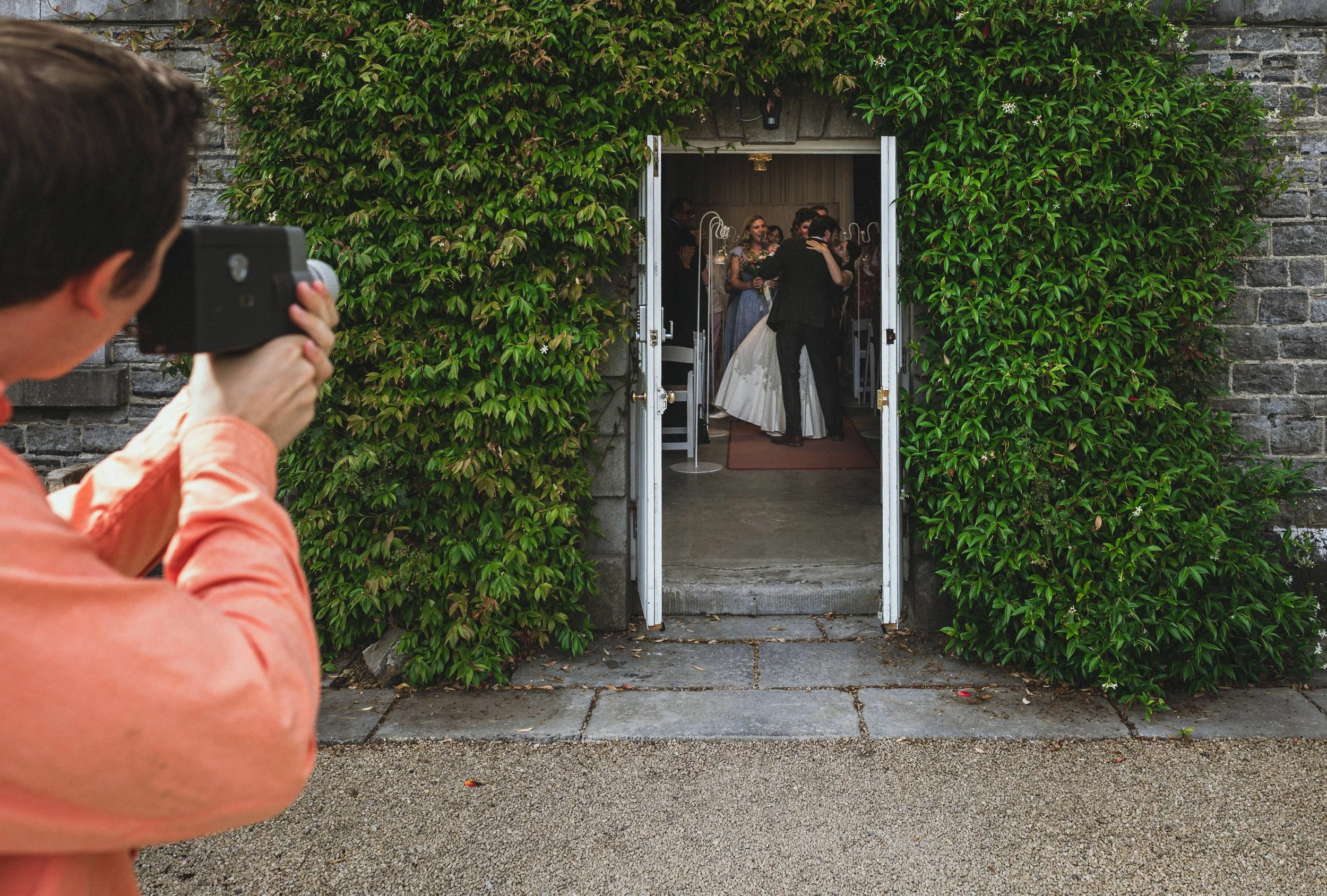 Person in orange shirt taking photo of wedding reception inside a building with green leafy exterior walls.