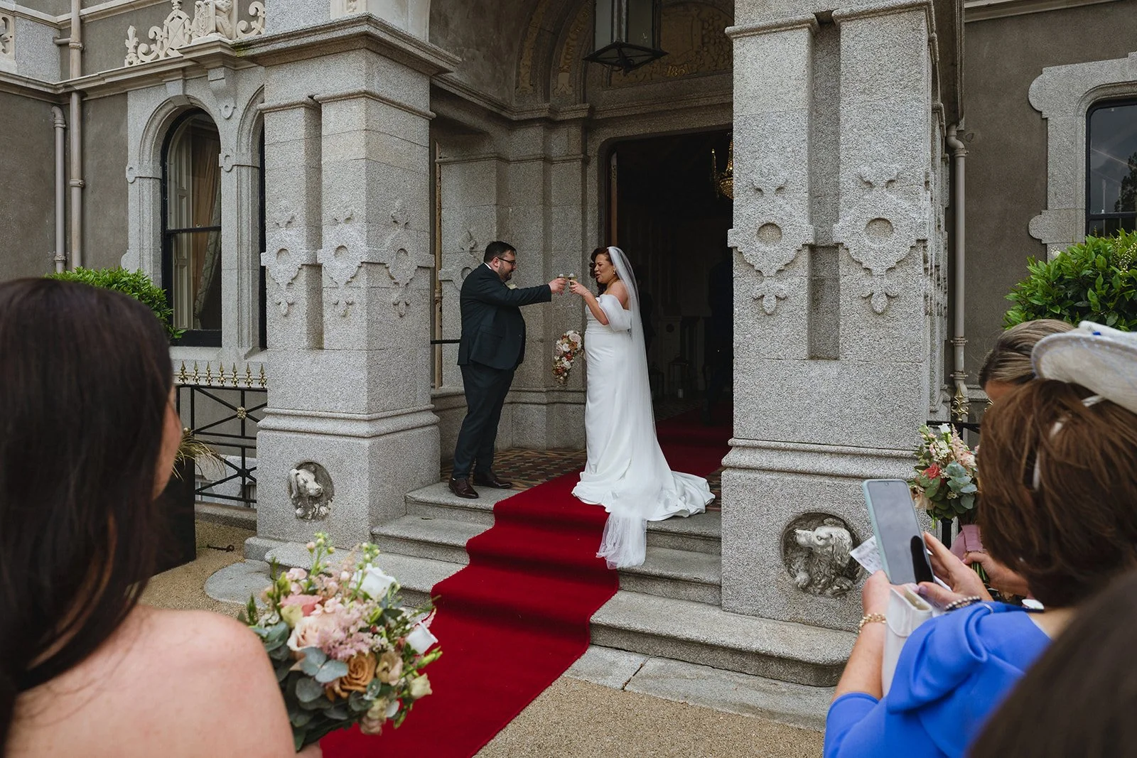 A bride and groom are standing on the steps of a building, exchanging a toast during their wedding ceremony. The bride is in a white wedding dress, and the groom is in a black suit. There are guests around, some taking photos, and floral arrangements are visible.