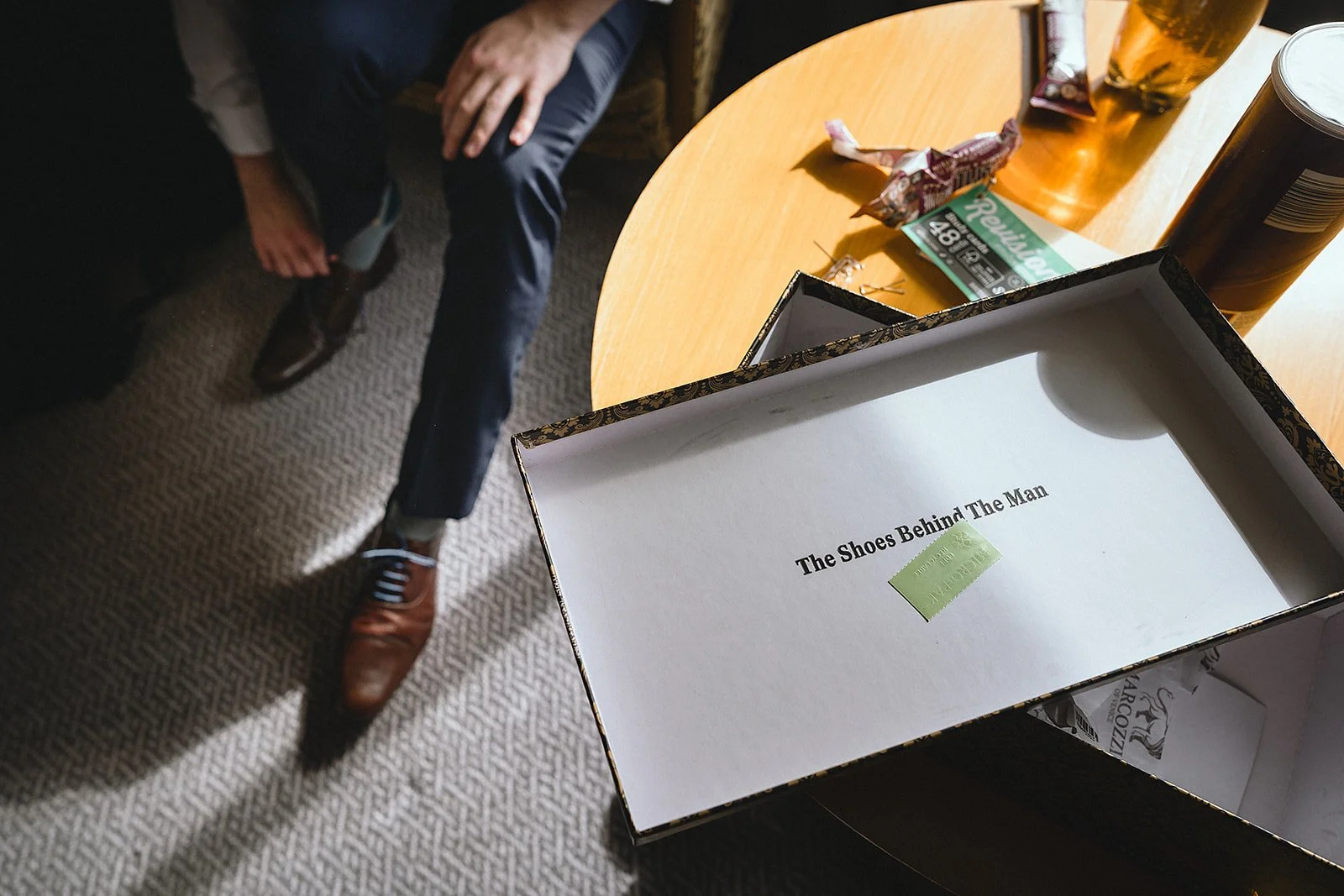 Open shoebox on a table with the title "The Shoes Behind The Man" and a green sticker. A man in study's dark pants and brown shoes is sitting nearby. The table has drinks, snacks, and a lottery ticket on it.