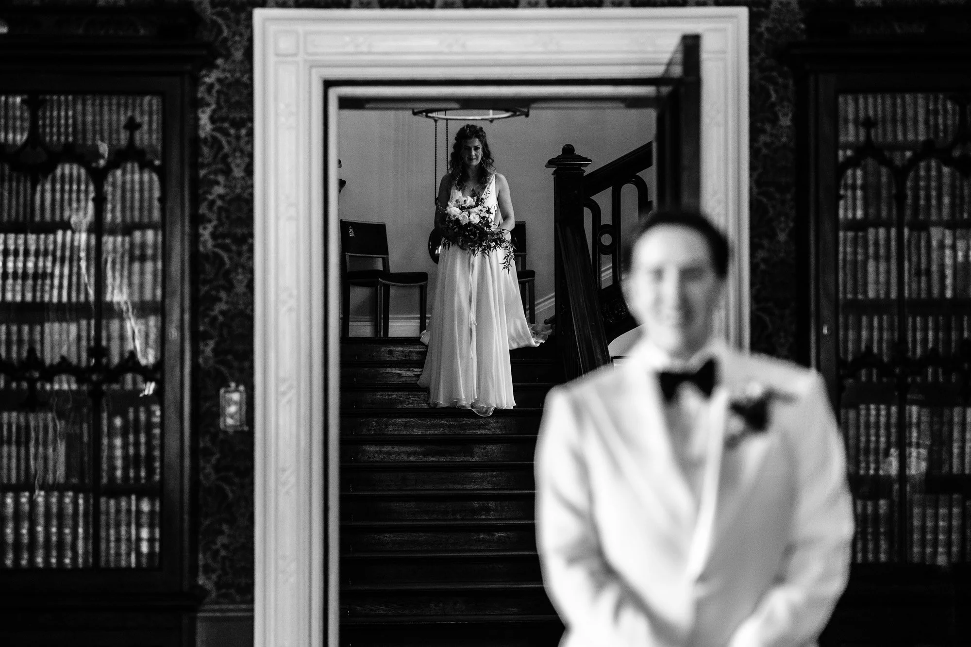 A woman in a white dress holding a bouquet of flowers, seen through a mirror, with a man in a tuxedo blurred in the foreground.