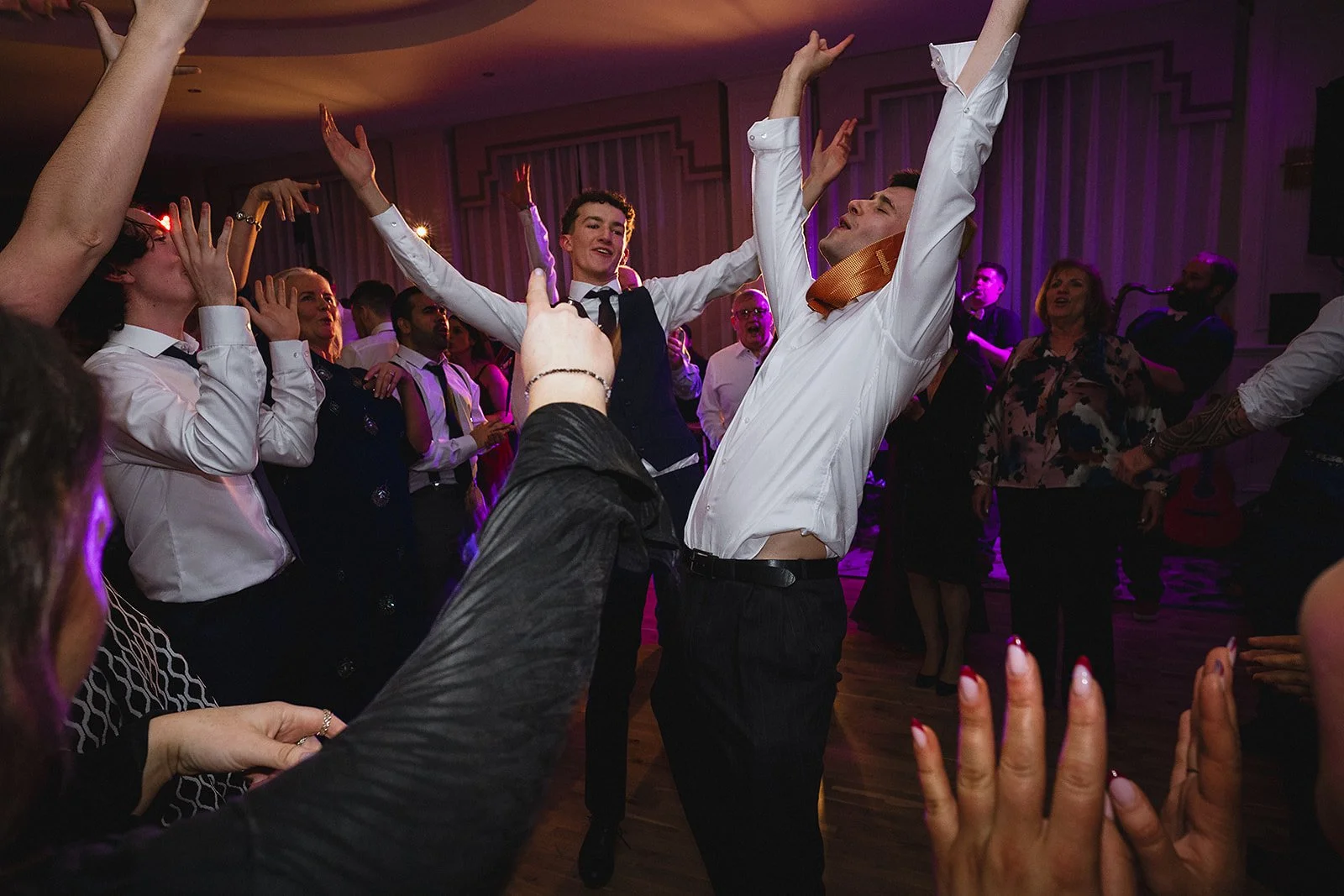 People dancing and celebrating at a party or wedding reception with purple lighting, some with hands raised and some smiling.
