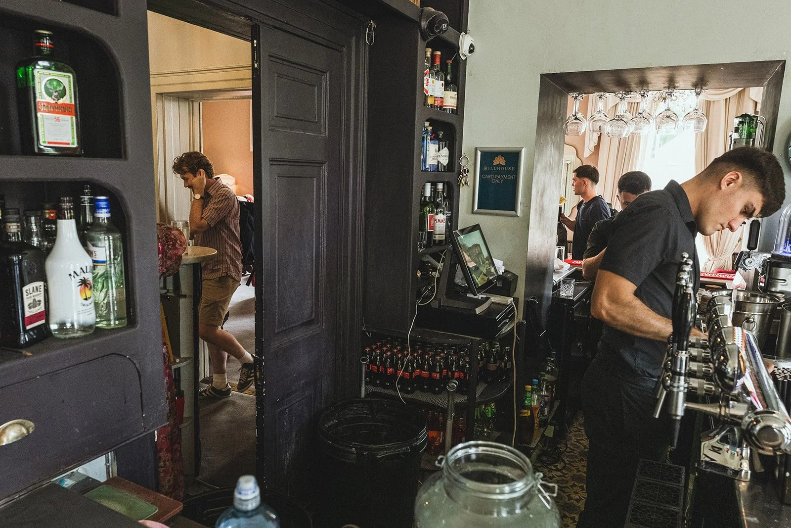 A bartender stands behind a bar counter preparing drinks, with three men in dark clothing working at a bar area in the background. A man in a plaid shirt and shorts is visible through a doorway, talking on the phone. Shelves with bottles and glasses 