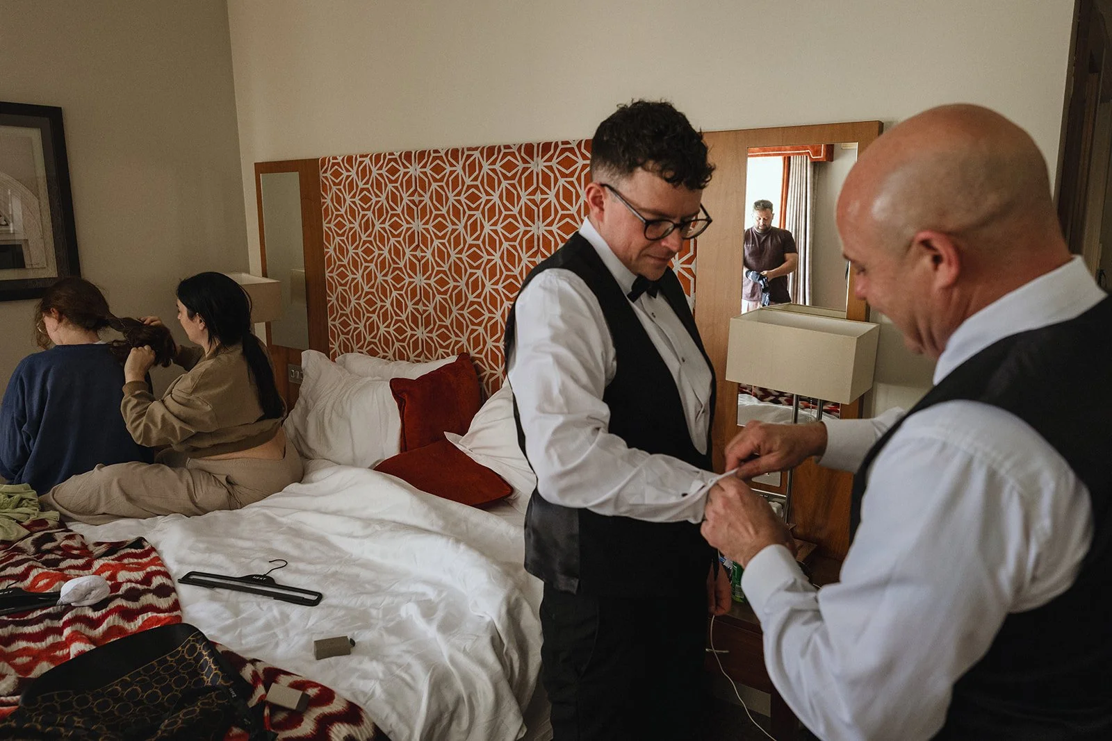 Two men in tuxedos assisting each other with a wardrobe in a hotel room, two women sitting on the bed, one with long dark hair, the other with curly brown hair, and a person in the doorway.