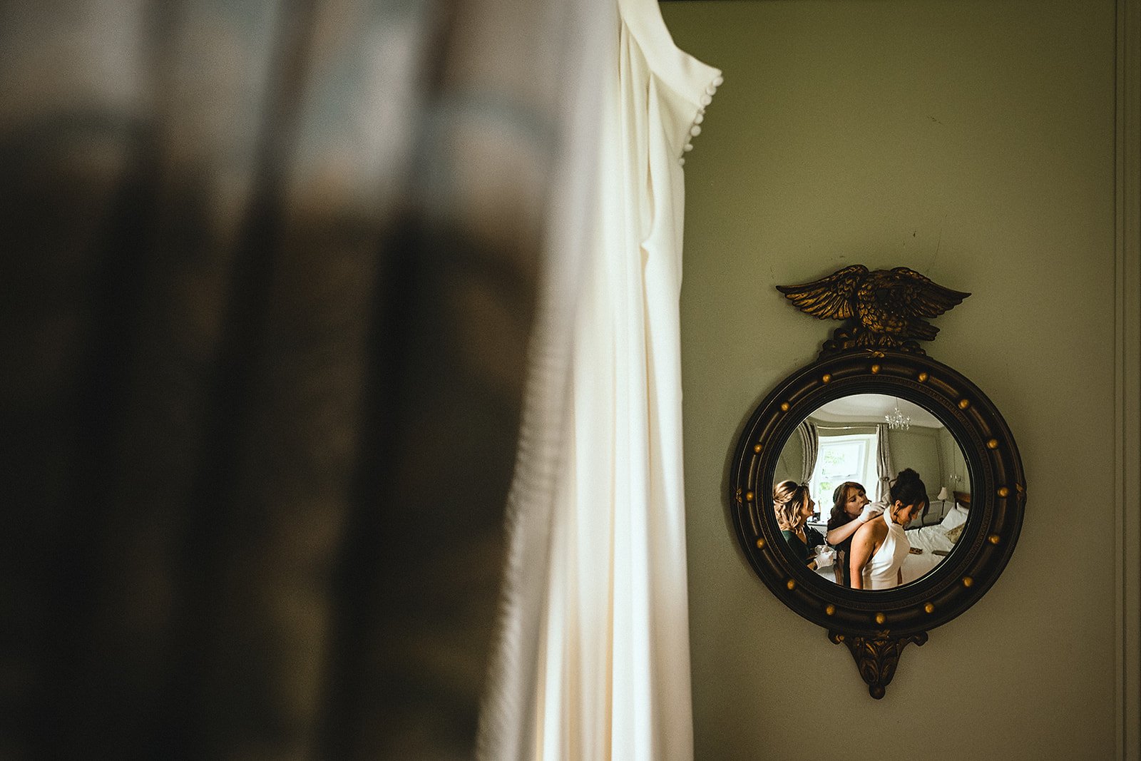 A round mirror with gold accents on a green wall, reflecting three women in a room, with one woman in a white dress having her hair styled by another woman.
