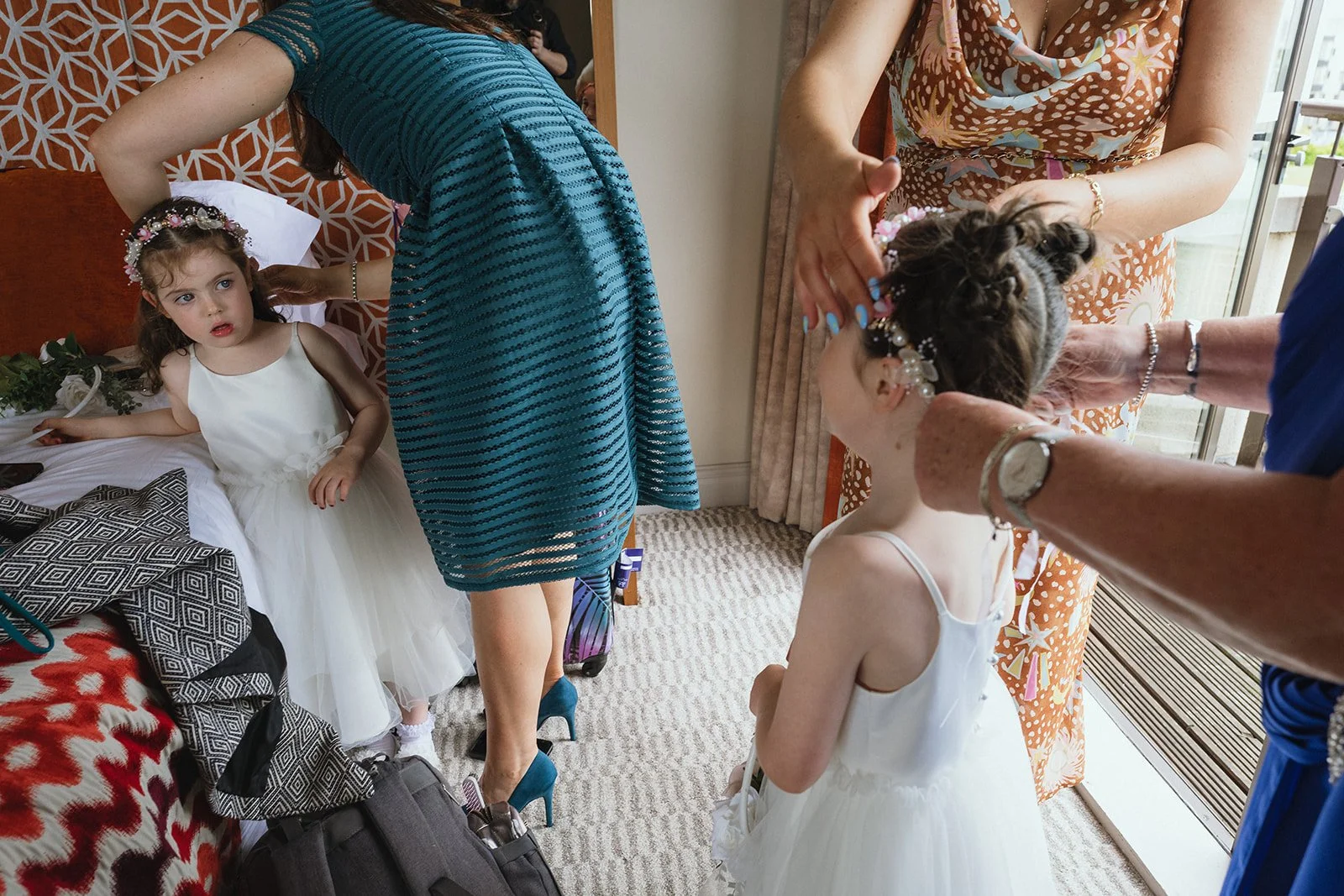 A young girl in a white dress with a floral headband sitting on a bed, looking surprised or curious, with other women helping two girls dress up, in a room with natural light and a balcony outside.
