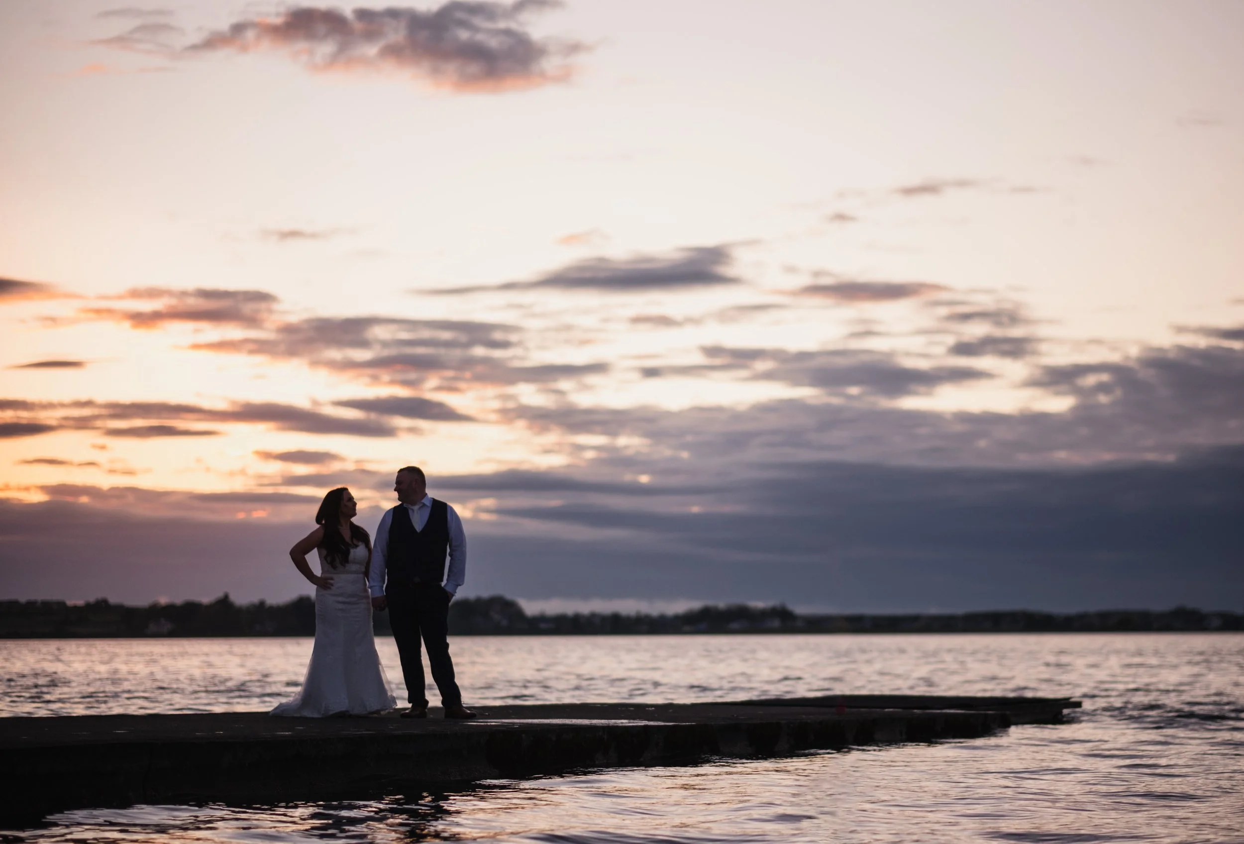 A couple dressed in formal clothing stands by a body of water during sunset, with clouds in the sky and the horizon in the background.