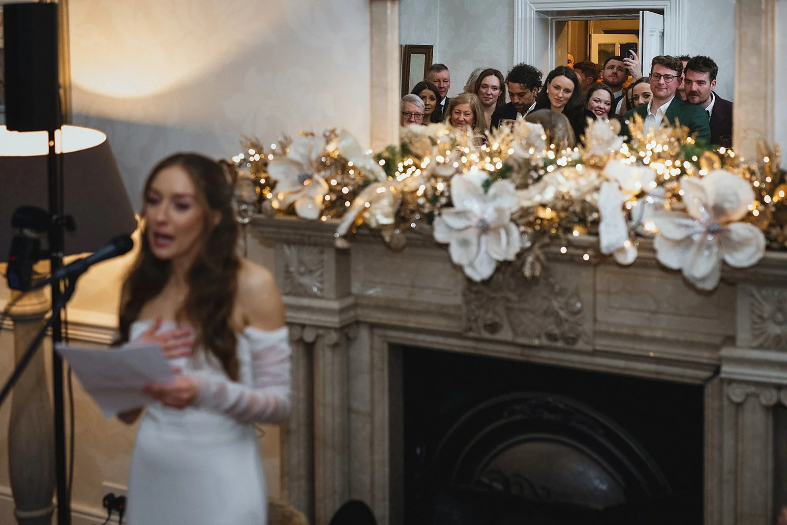 A woman in a white dress reading from a paper at a microphone, in front of a fireplace decorated with white flowers, ribbons, and string lights, with a crowd of people watching and smiling from behind the fireplace.