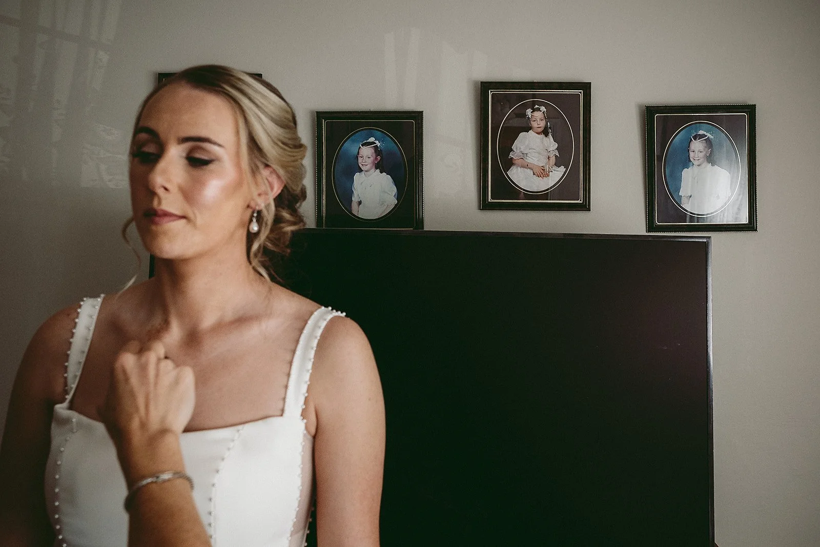 A woman in a wedding dress standing in a room with three framed childhood photos hanging on the wall behind her.
