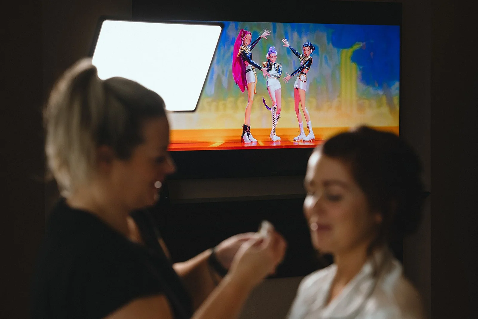 Two women are in a makeup or hair styling session in front of a colorful poster of three animated characters, possibly in a theatrical or promotional setting.