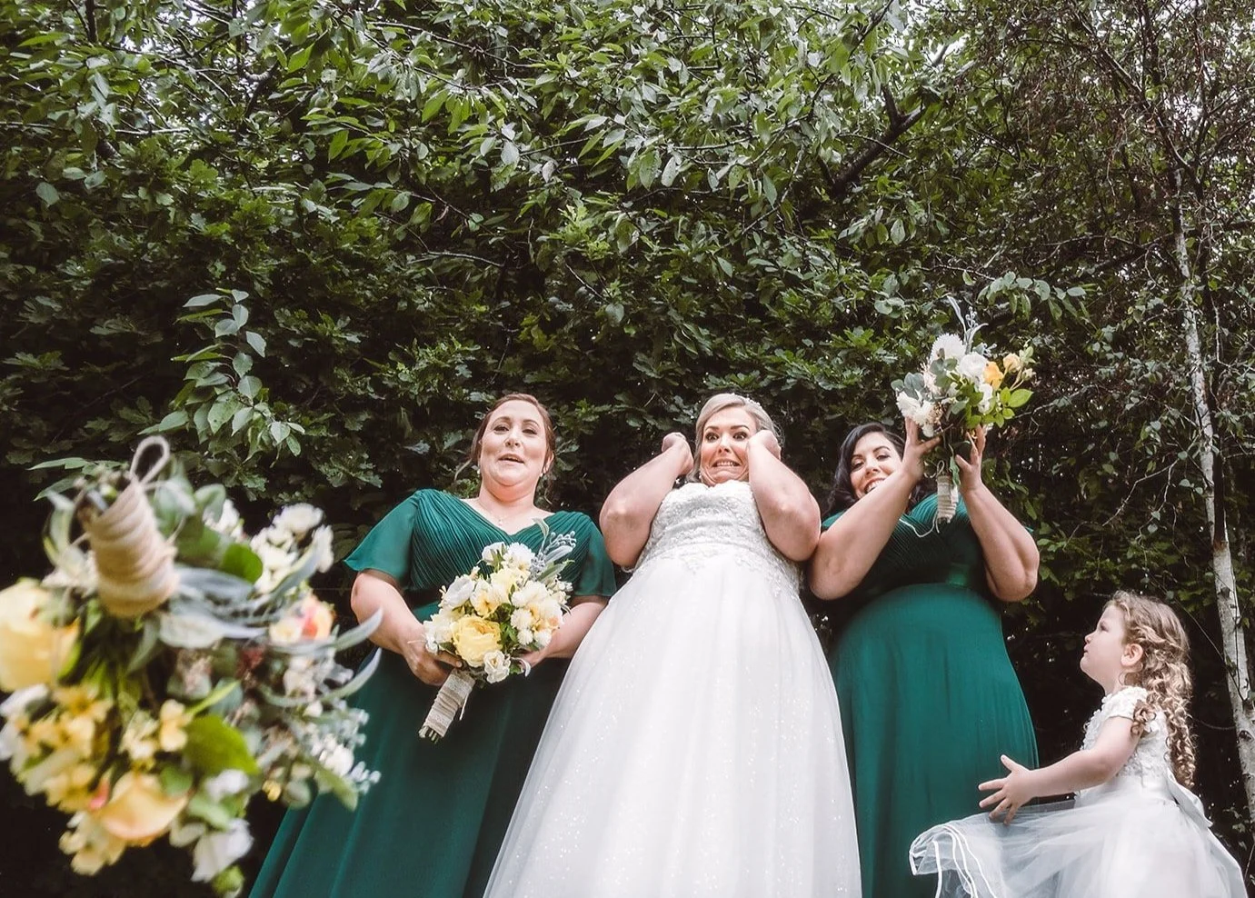 Bride and bridesmaids in green dresses with bouquets standing outdoors with trees in the background, humorous expressions.