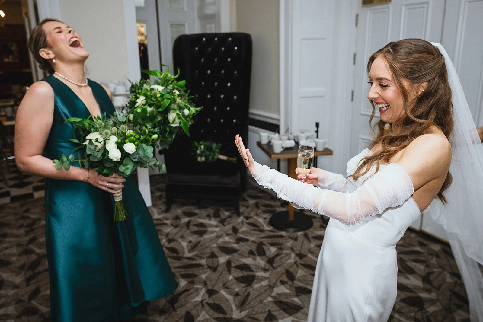 A joyful bride in a white wedding dress, holding a glass of champagne, is smiling and raising her hand towards a woman in a teal dress holding a bouquet of white and green flowers, all indoors in a decorated room.