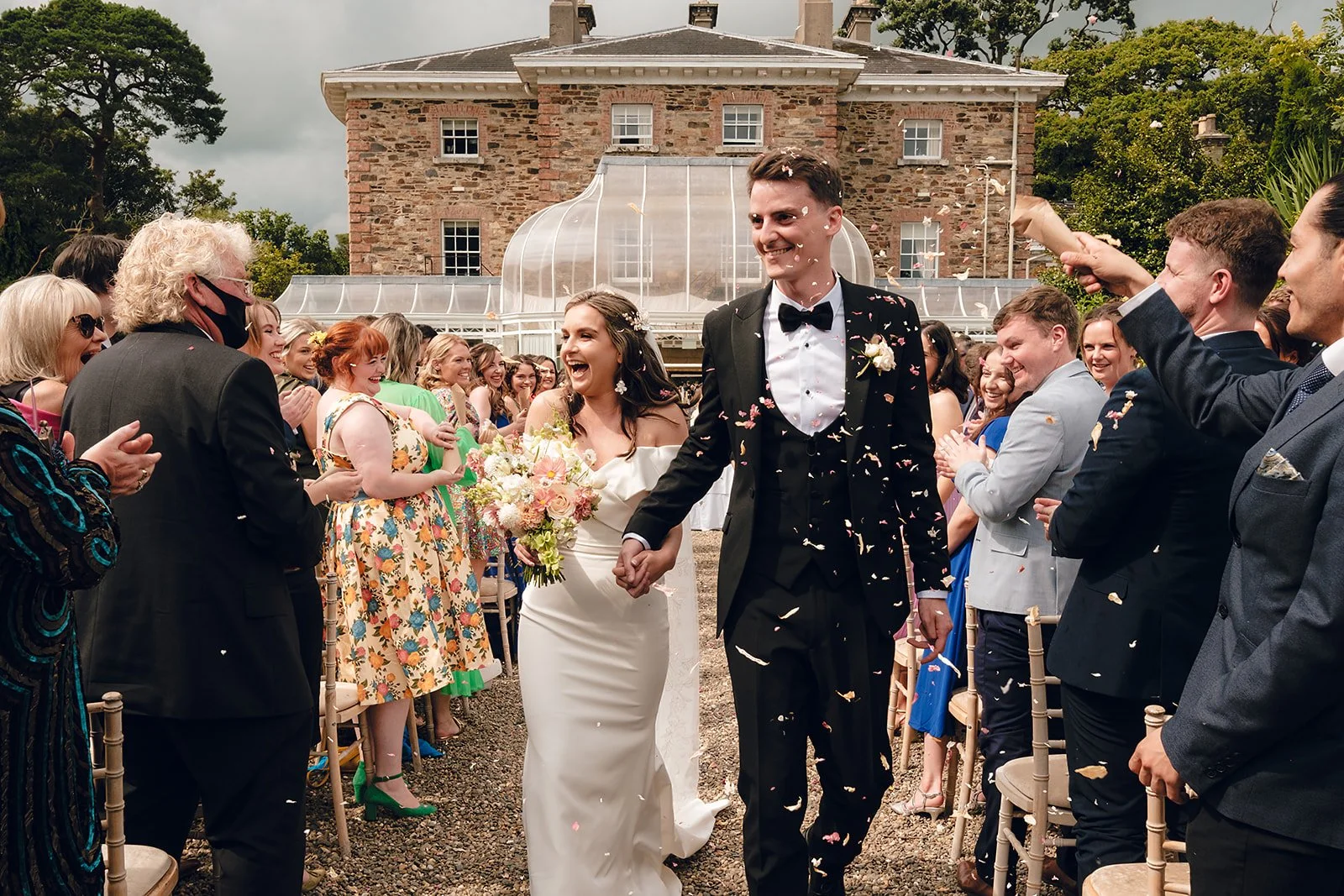 A newlywed couple walking hand in hand through a crowd of joyful wedding guests, with confetti falling around them, outside a rustic brick building with glass greenhouse extension.