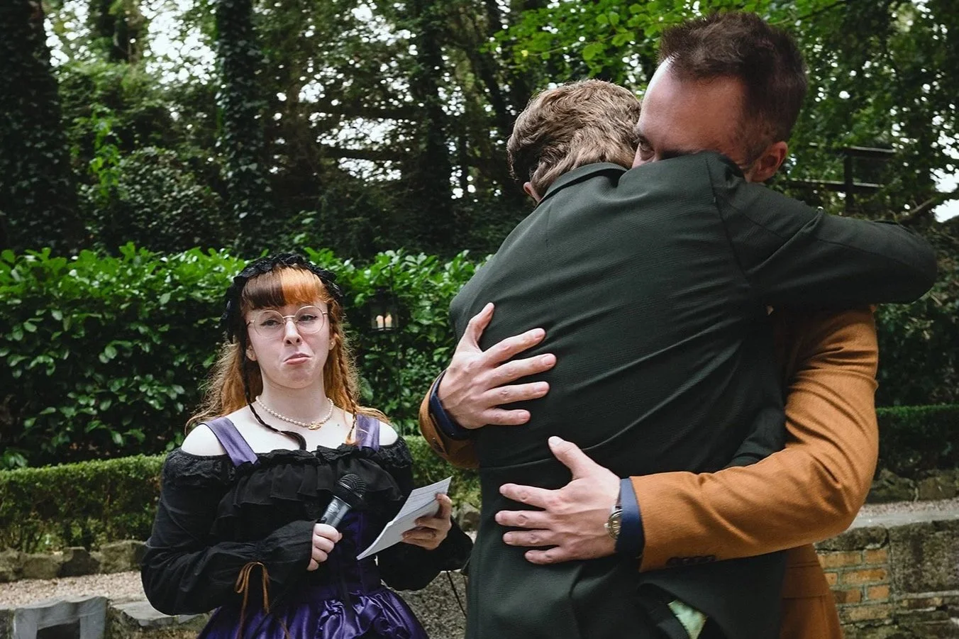 A young woman dressed in a Gothic style outfit holding a microphone and a note while watching two men hugging outdoors.