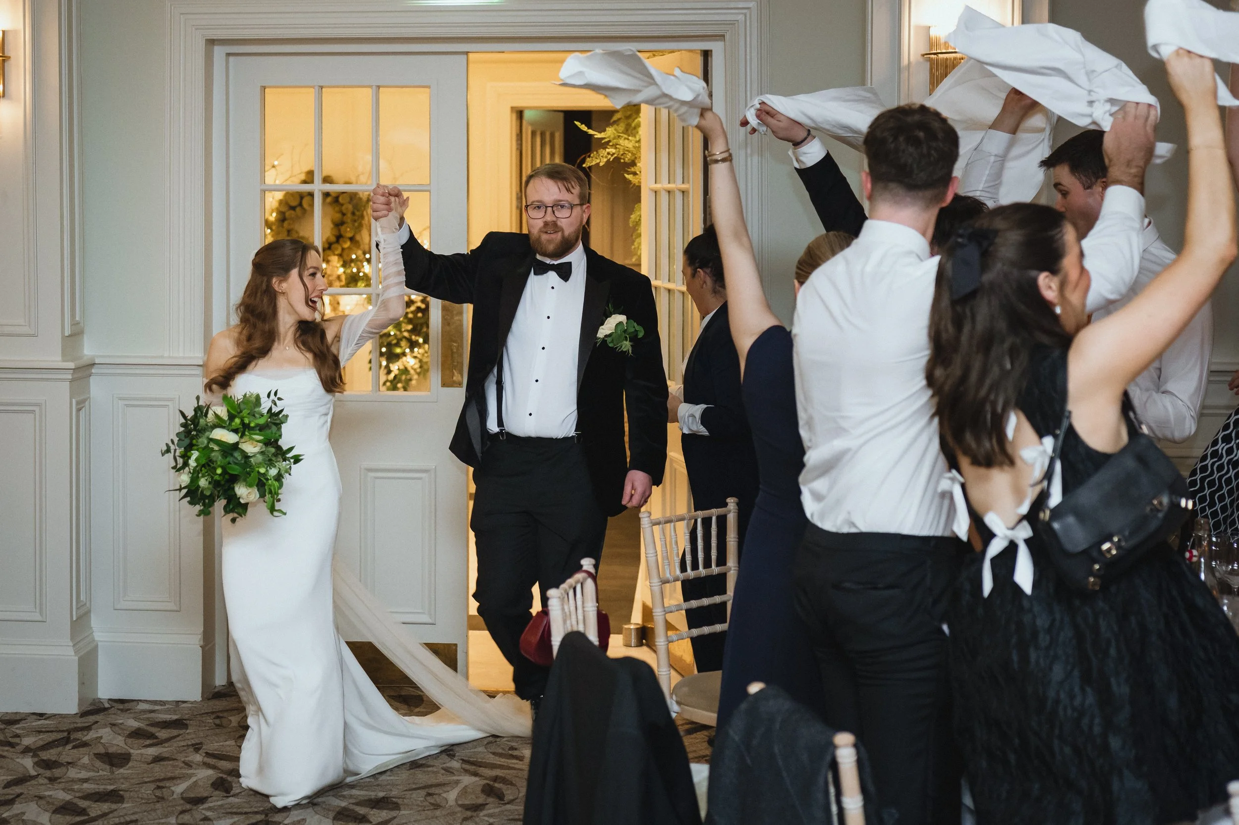 Bride and groom entering wedding reception with guests raising napkins in celebration.
