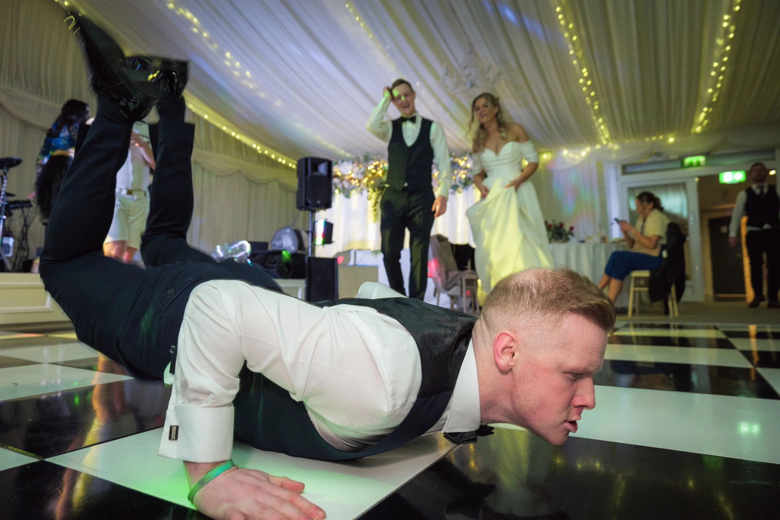 Man doing a push-up on a checkered dance floor at a wedding reception, with people and decorations in the background.