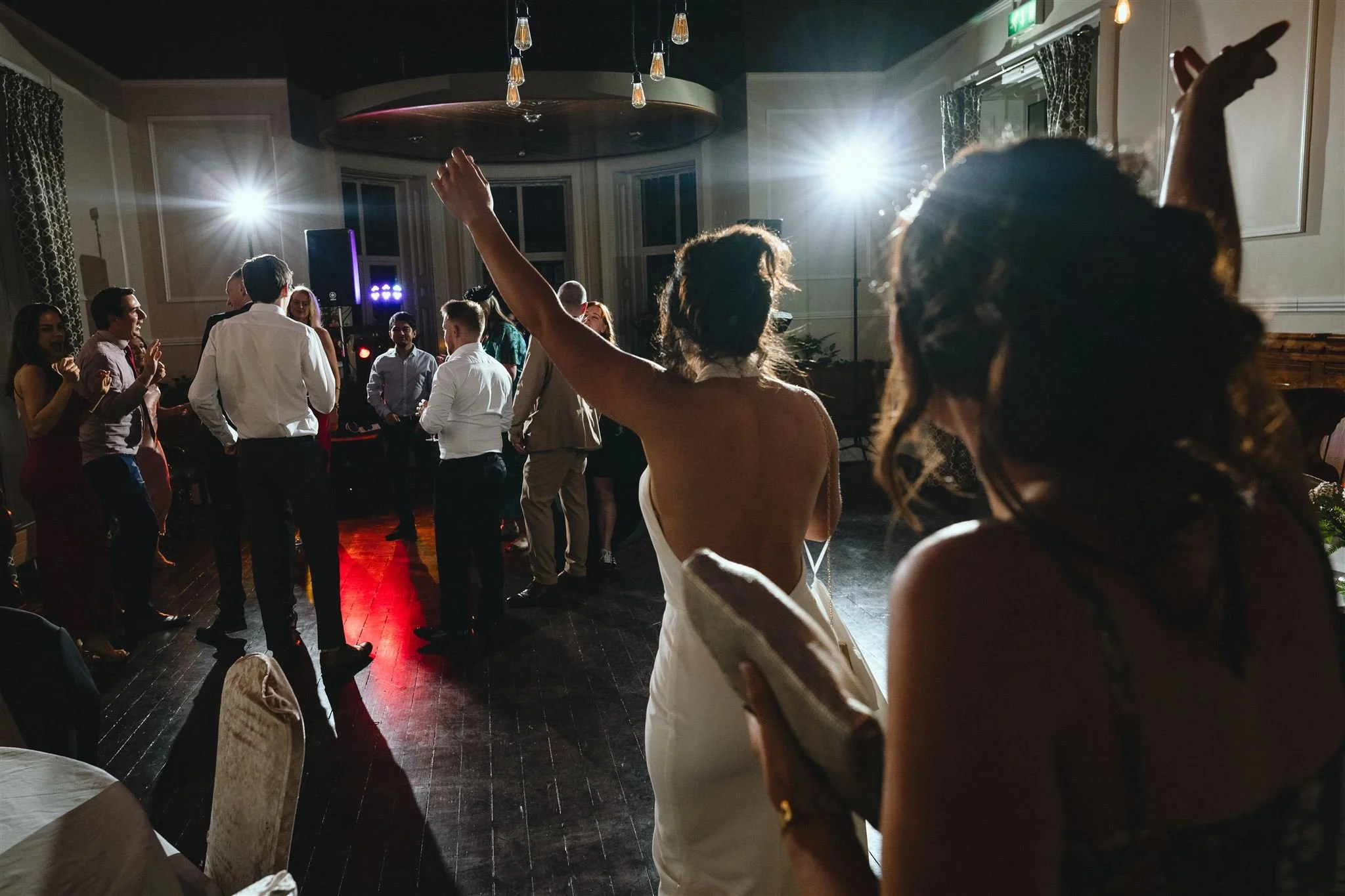 People dancing and socializing at a party or wedding reception in a large, elegant room with high ceilings and a dance floor.