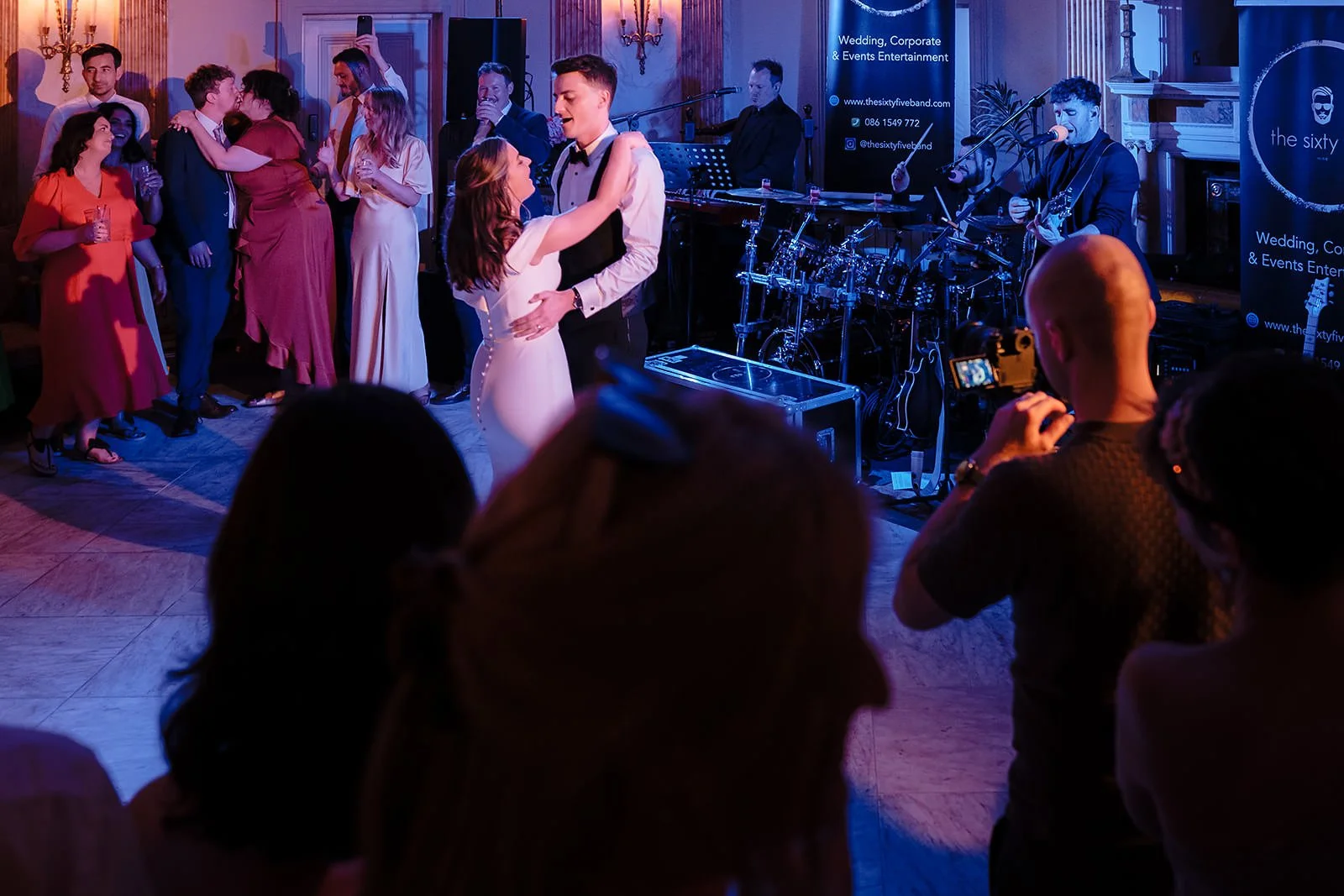 Couple dancing in the center of a crowded dance floor at a wedding reception, with a live band playing on stage in the background. Guests are watching and taking photos in a luxurious, dimly lit ballroom.