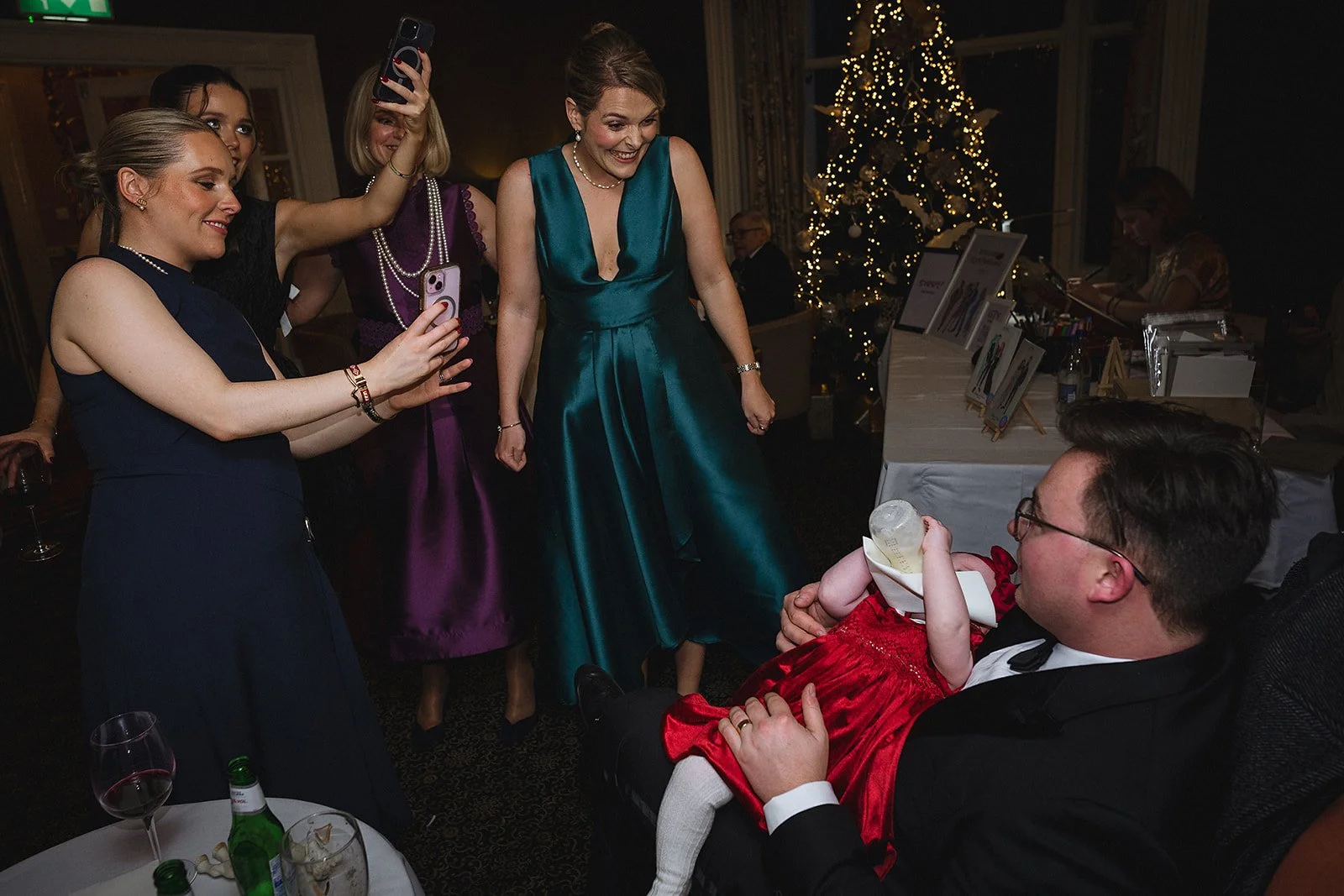Group of women and a man at a Christmas party, with a Christmas tree in the background. One woman is taking a picture of a man holding a baby dressed in a red dress. The man is wearing a tuxedo and glasses, and the women are dressed in formal evening