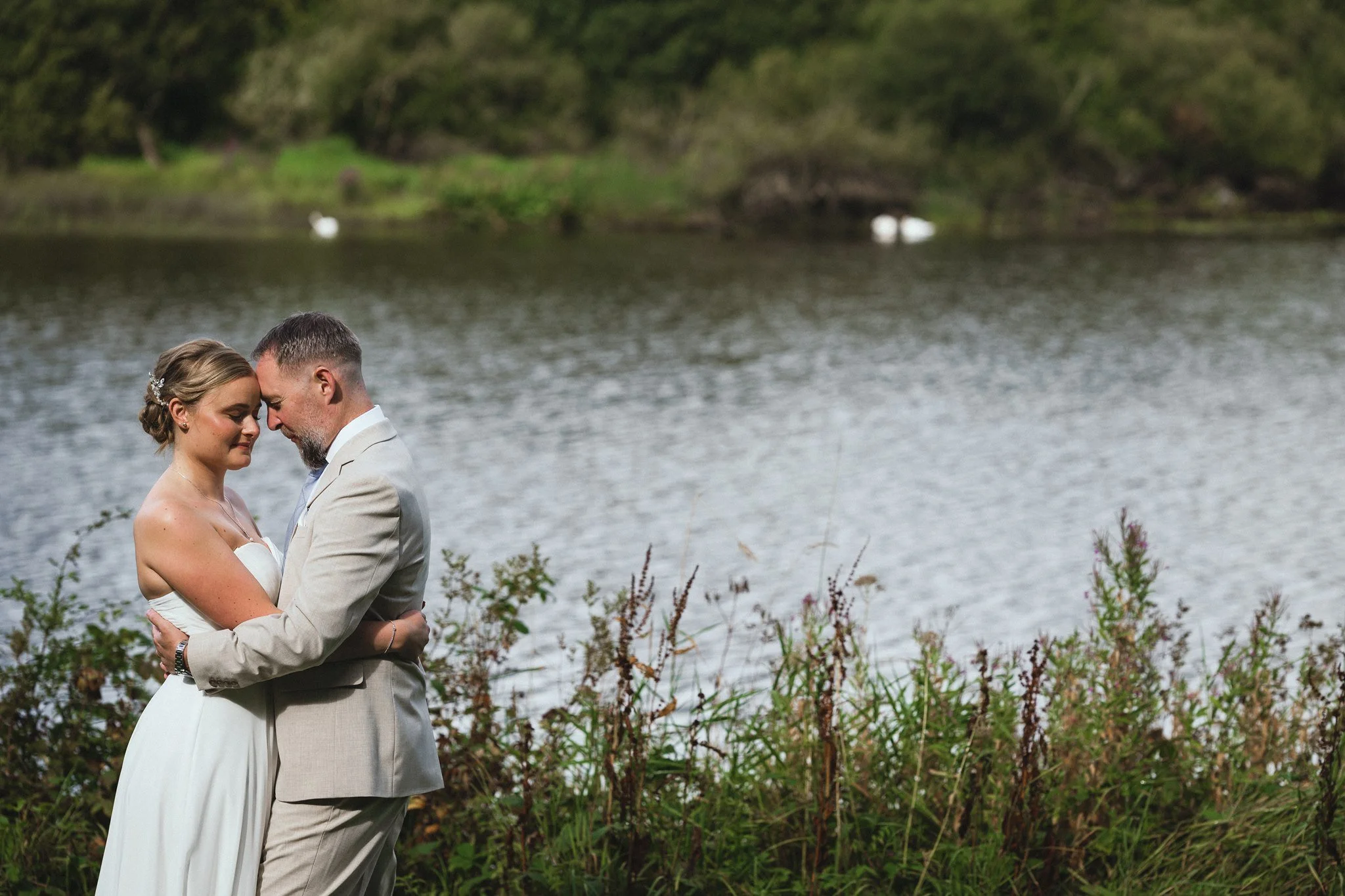 A bride and groom standing close together, touching foreheads and smiling by a lakeshore surrounded by green foliage, with swans on the lake in the background.