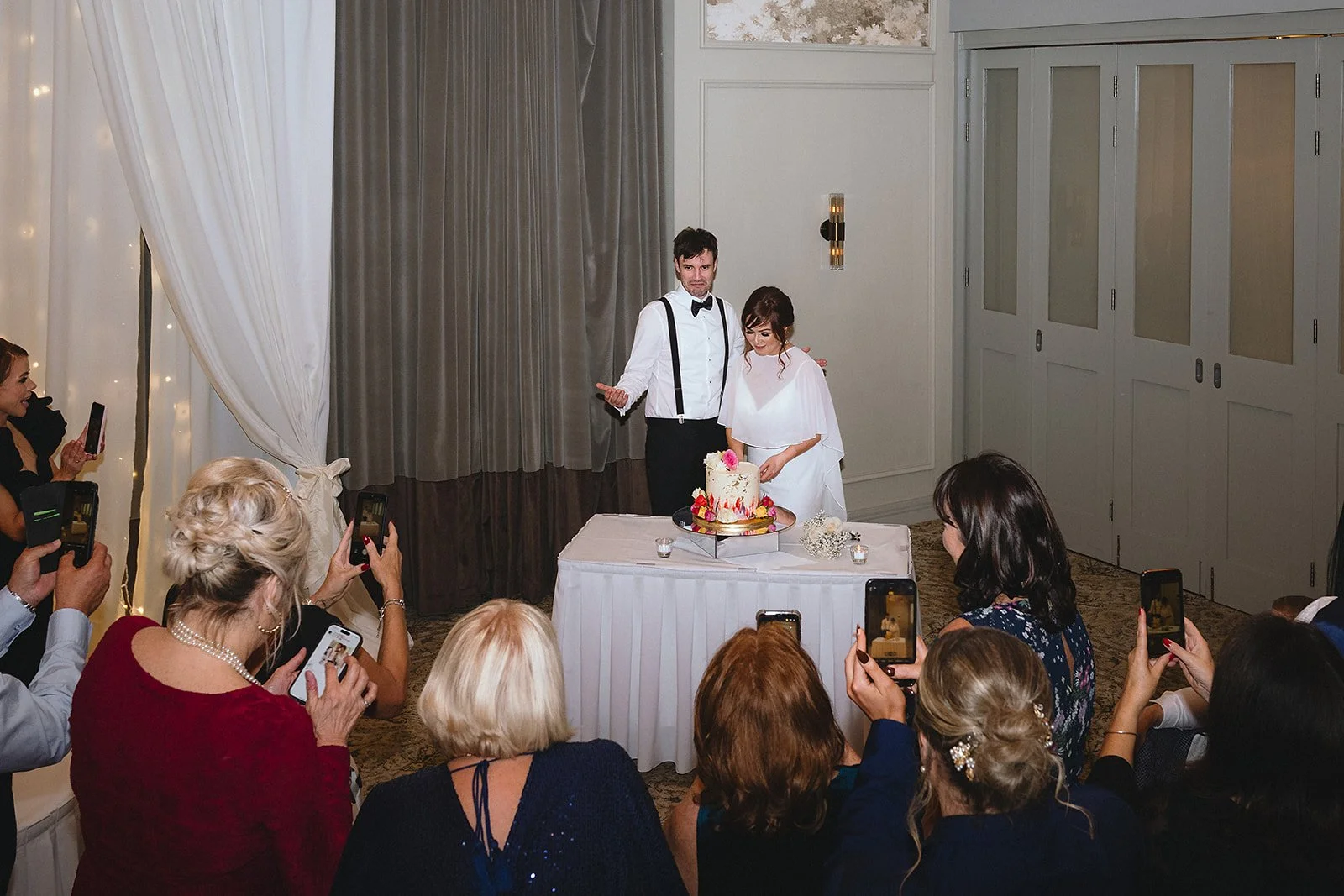 A bride and groom cutting a wedding cake at their reception, surrounded by guests taking photos.