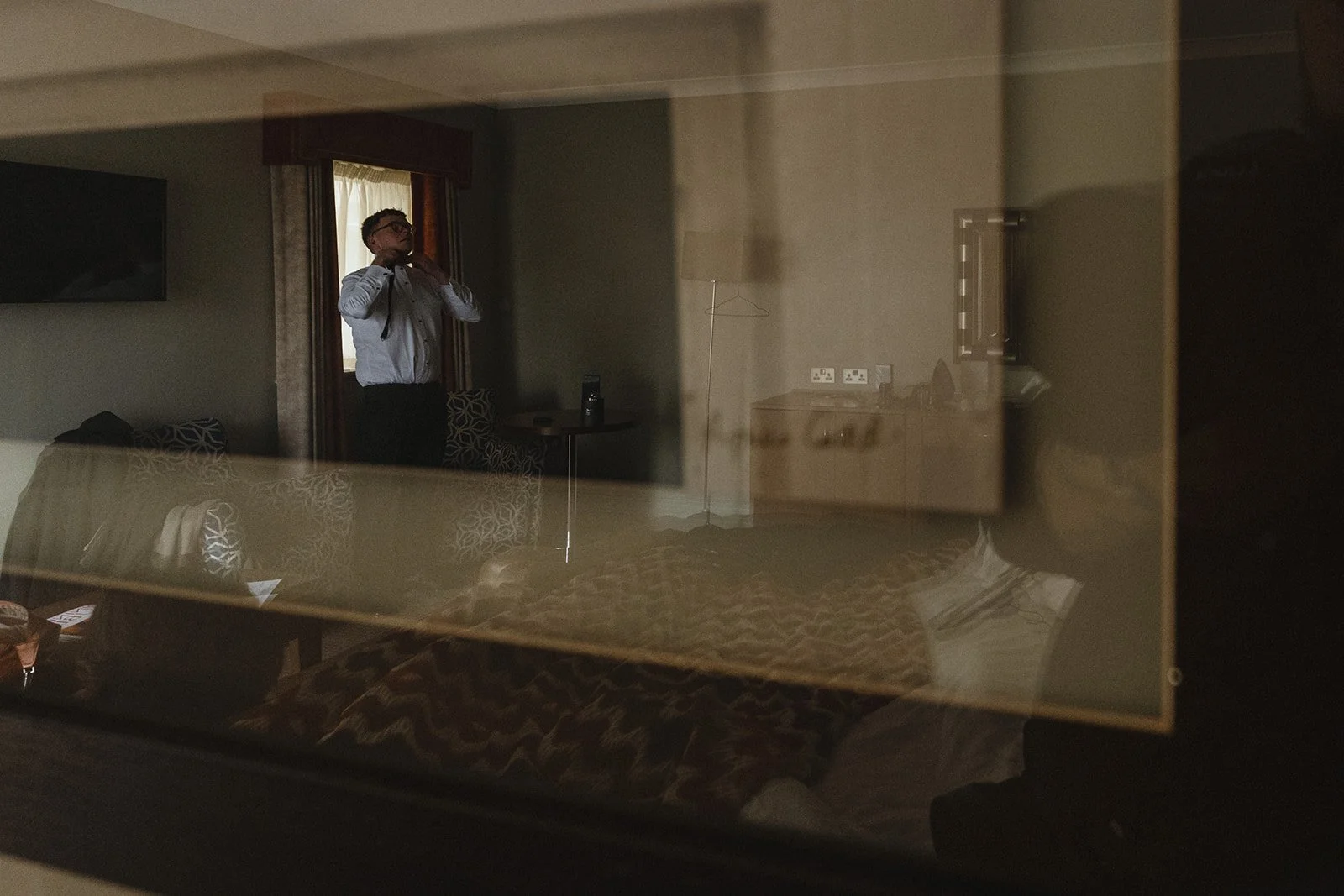 A man in a white shirt and glasses is adjusting his tie in a hotel room, seen through a reflective glass or mirror.